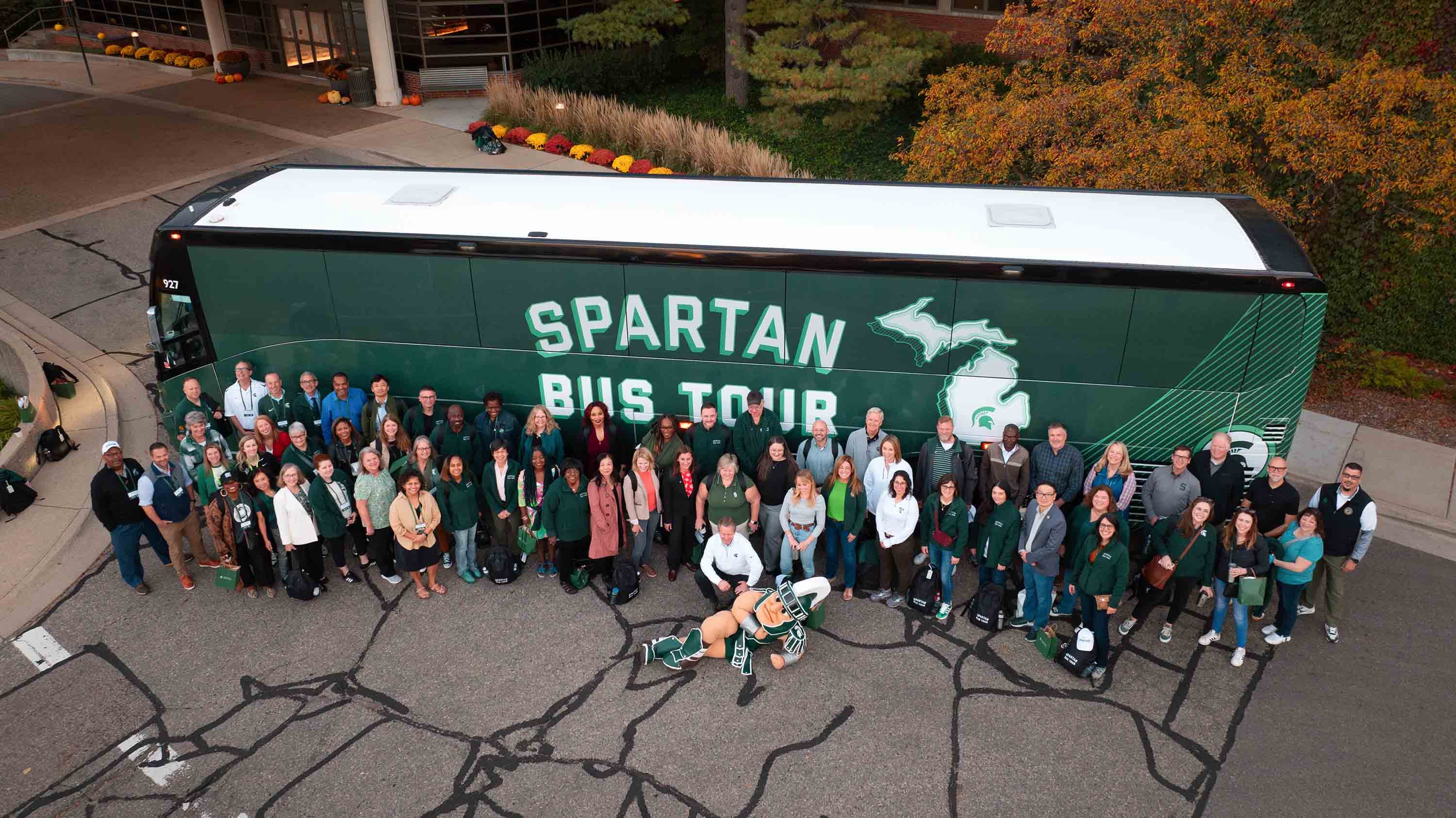 n aerial view of people posing for a group photo in front of a bus. The bus is green and decorated with the words ?Spartan Bus Tour? and has the outline of the state of Michigan with a Spartan helmet logo where East Lansing is located.