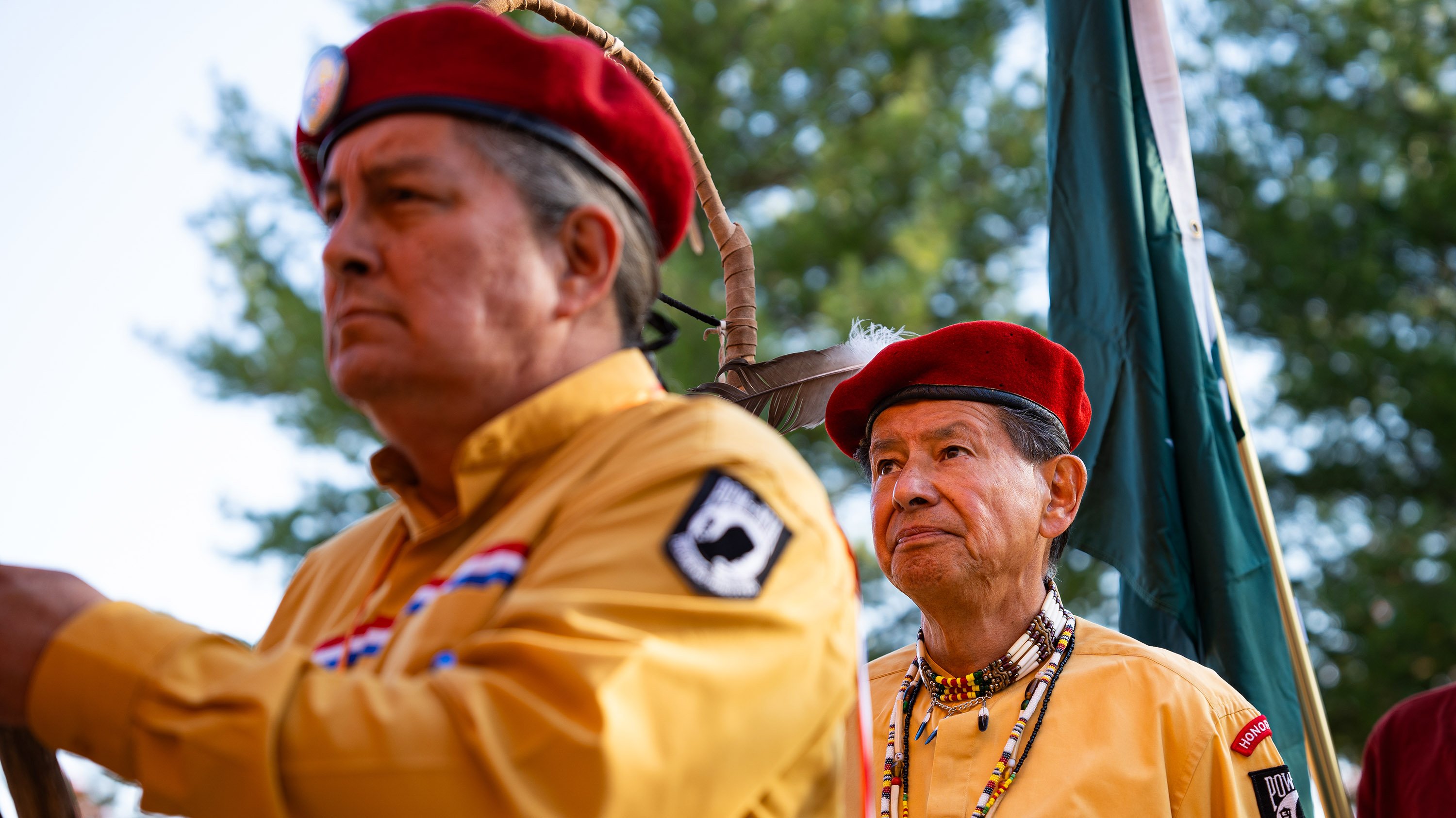 A close-up of men carrying flags. They are wearing red berets and mustard yellow shirts with a black and white POW-MIA patch. One of the men is wearing traditional Native American beads.