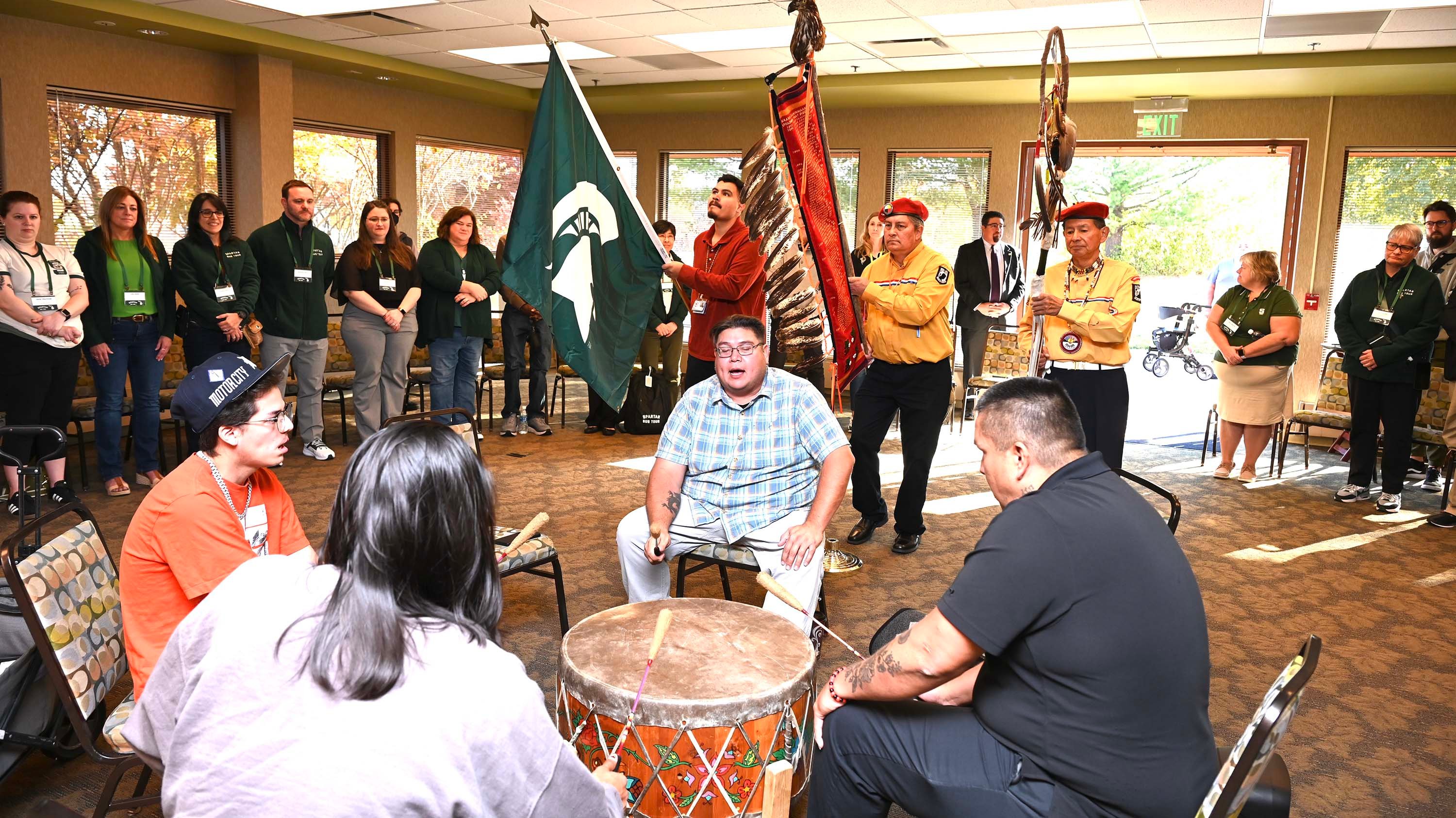Four people sit in a drum circle and play music with flagbearers behind them. A circle of people watch.
