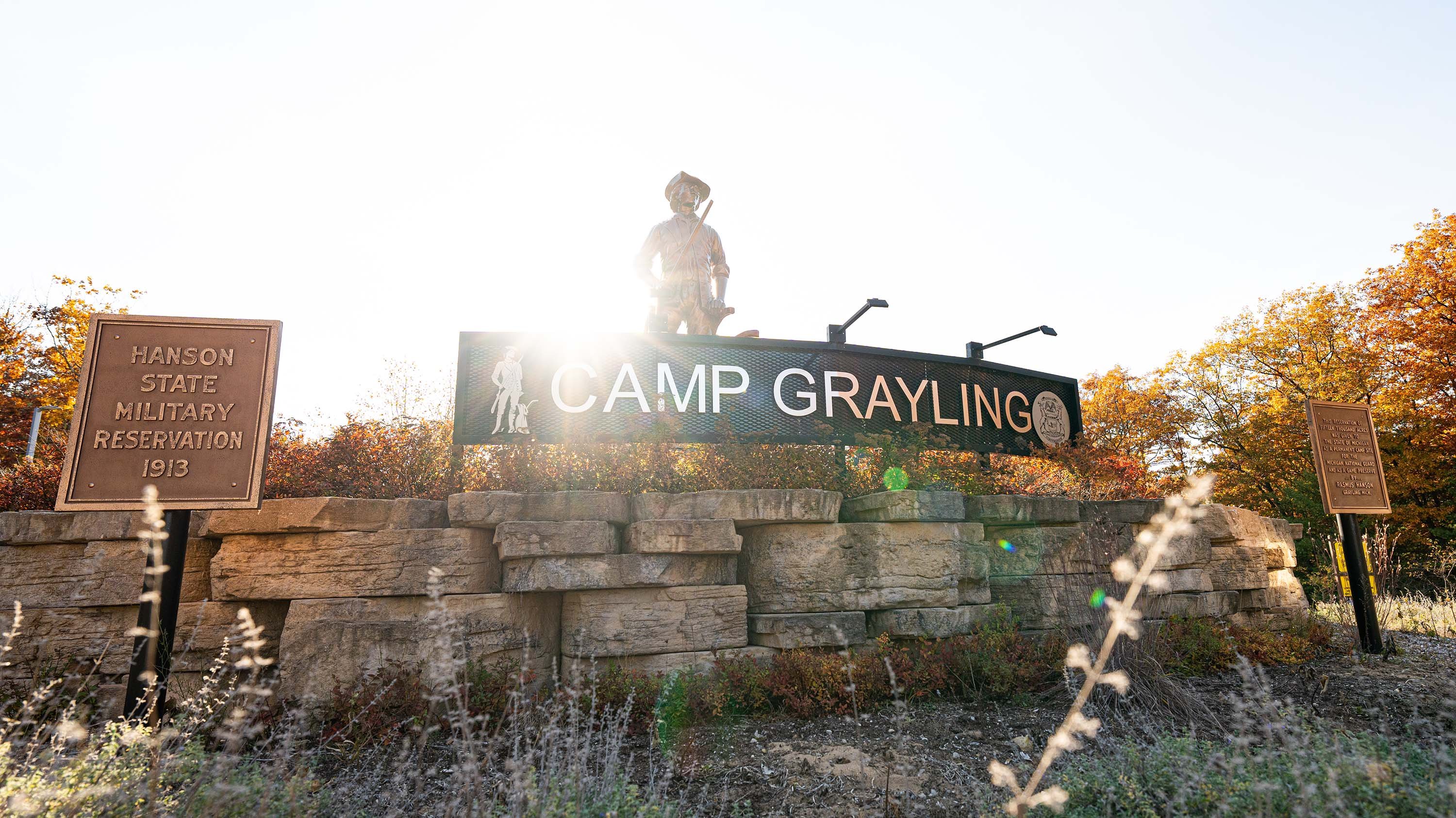 A metal sign that says ?Camp Grayling? rests on top of stone. There is a statue of a soldier behind the sign. Next to the sign, there is a bronze plaque that reads ?Hanson State Military Reservation 1913.?