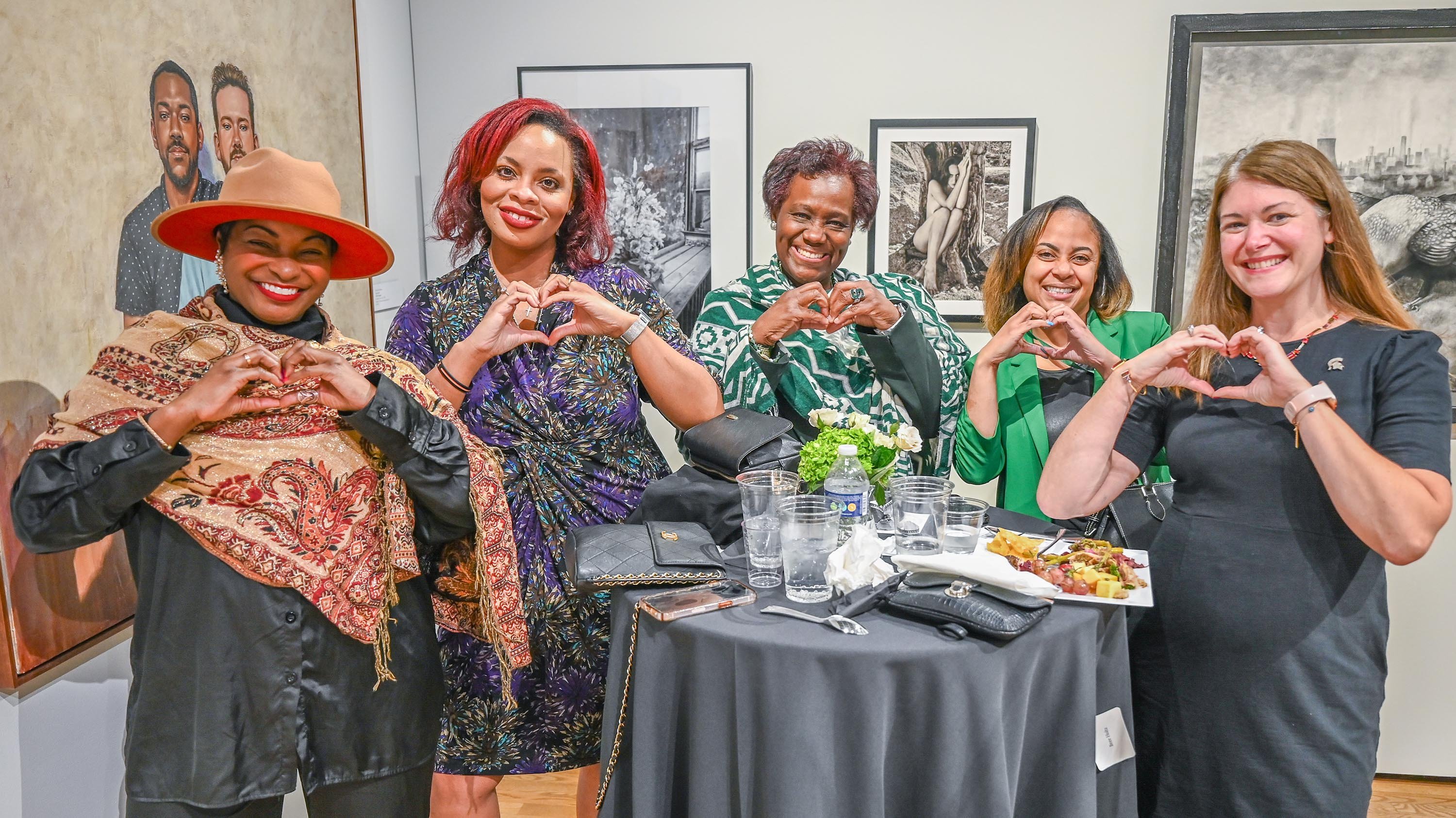 A group of people posing for a picture in an art gallery. They are standing around a table and making a heart shape with their hands.