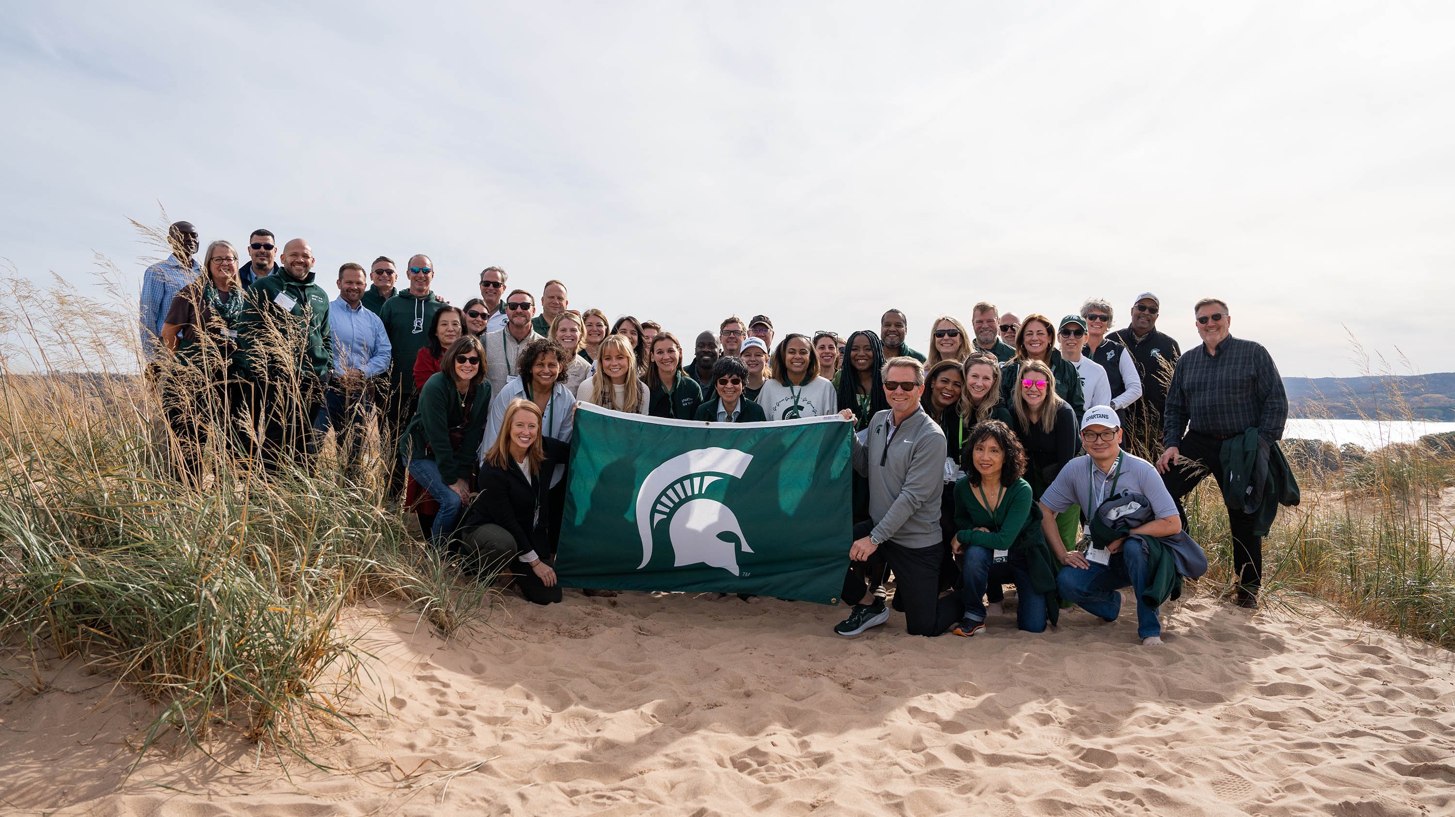 A group posing for a photo on a sand dune. Two people are holding the a green flag with a white Spartan helmet logo.