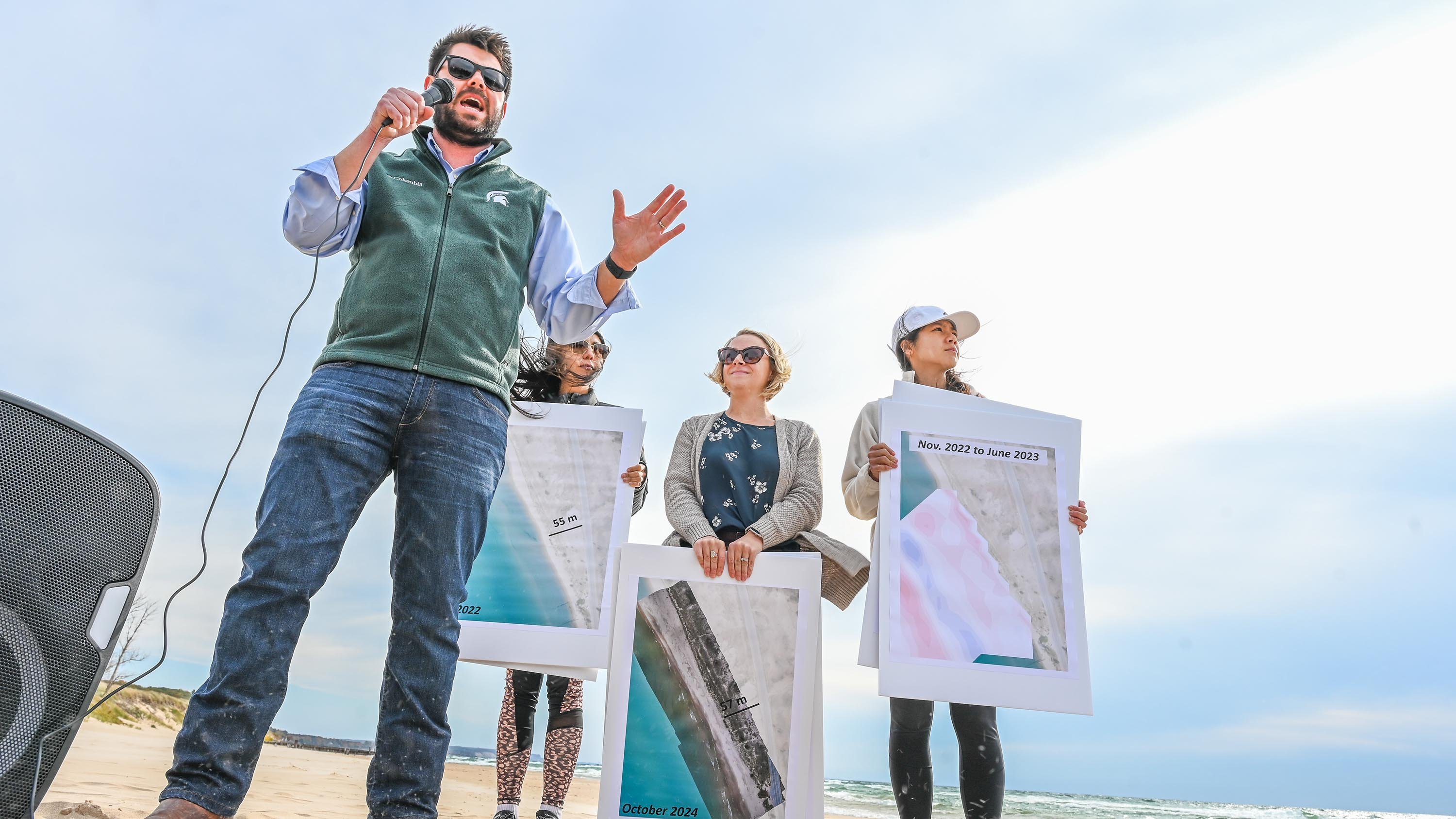 A man in talks into a microphone as three people holding posters stand behind him.