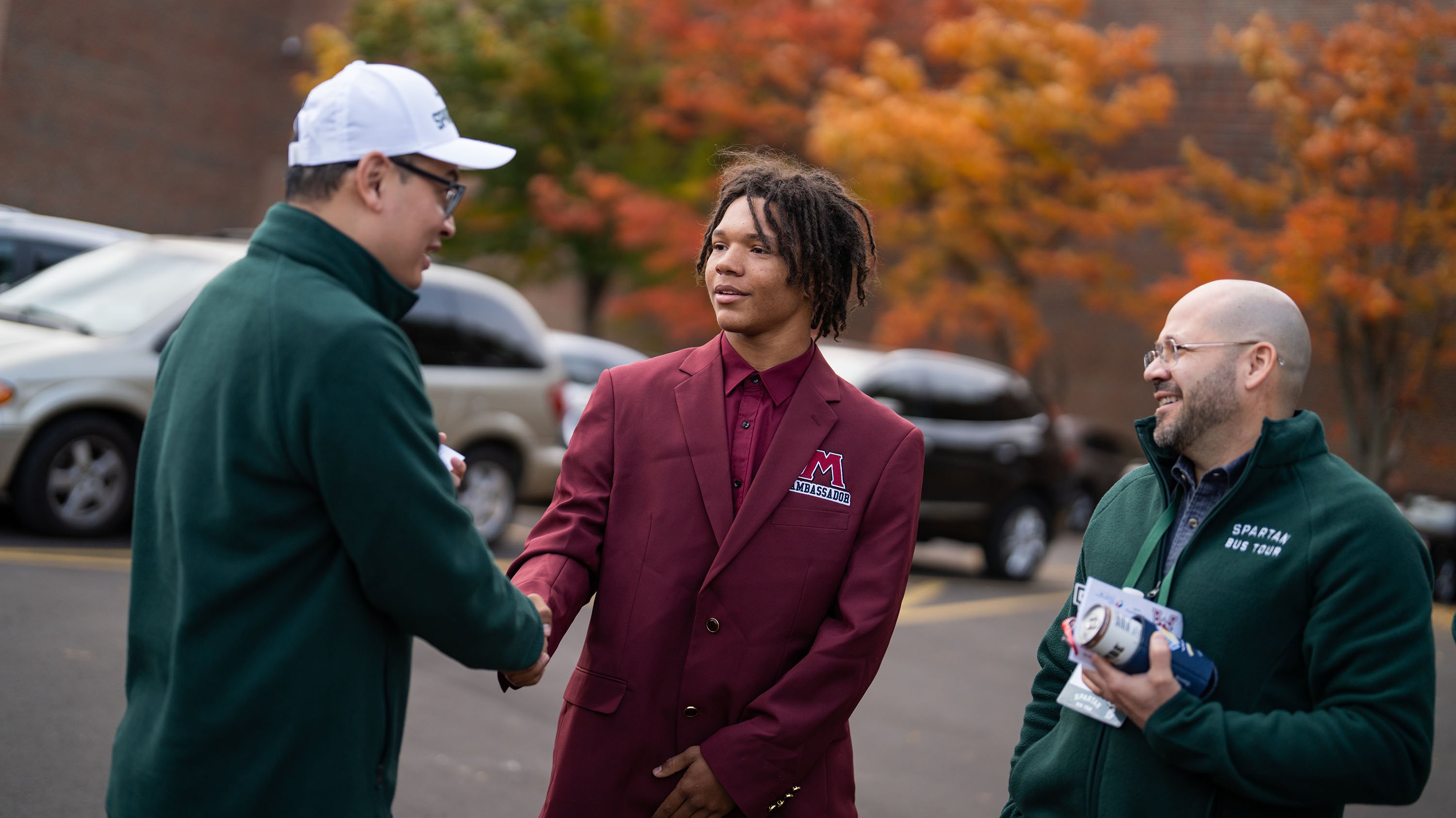 A person wearing a maroon blazer, dress shirt and tie with the Muskegon High School logo and text ?ambassador? underneath shakes hands with someone wearing a green fleece jacket and white hat as someone else in a green fleece jacket looks on.