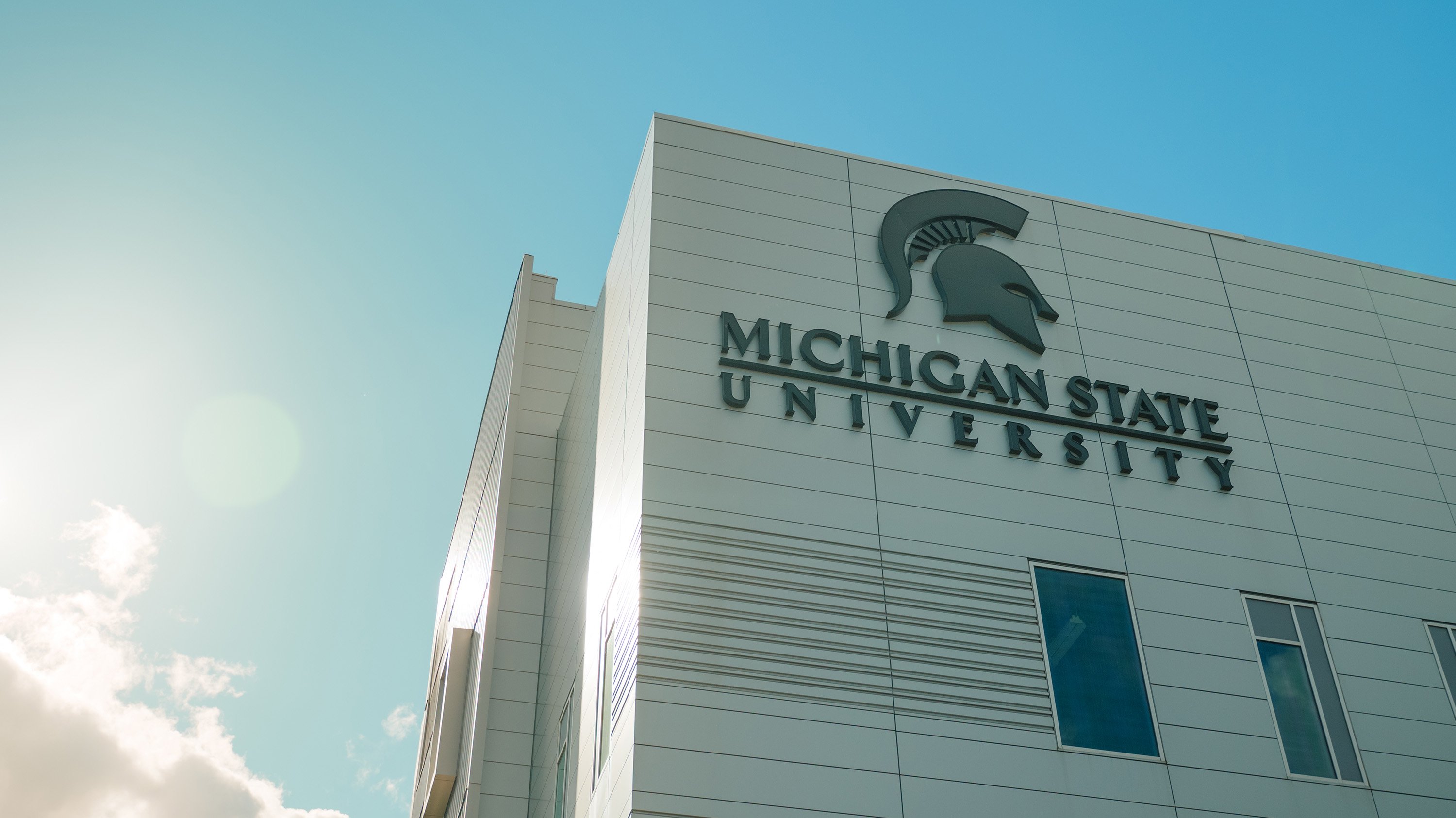 A green  Michigan State University wordmark and Spartan helmet logo at the top of a silver building.