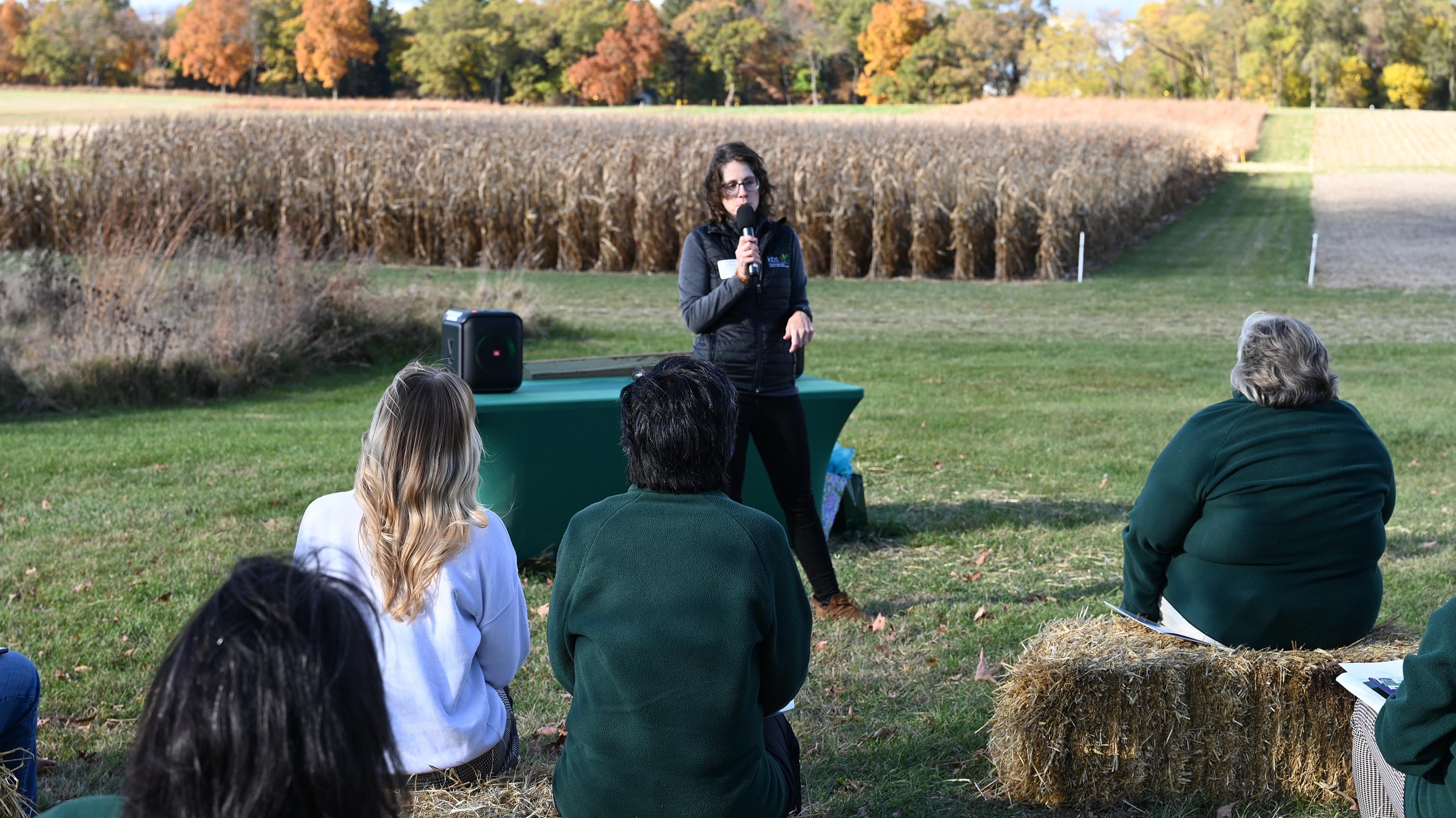 A person giving a presentation outdoors near a cornfield. The audience is sitting on bales of hay.