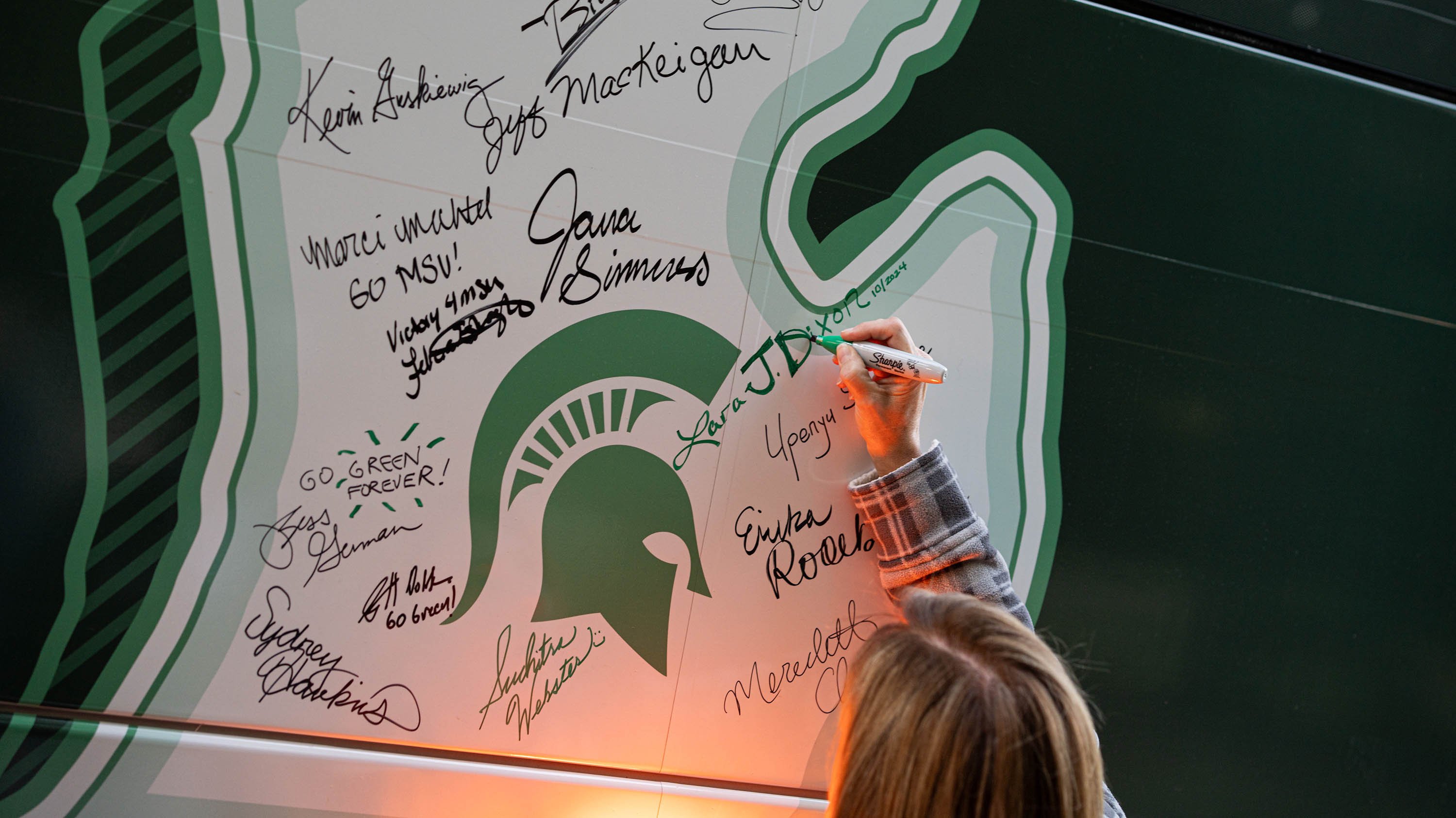 A person signs the white shape of the state of Michigan on a green bus with a green permanent marker. There are other signatures around theirs.