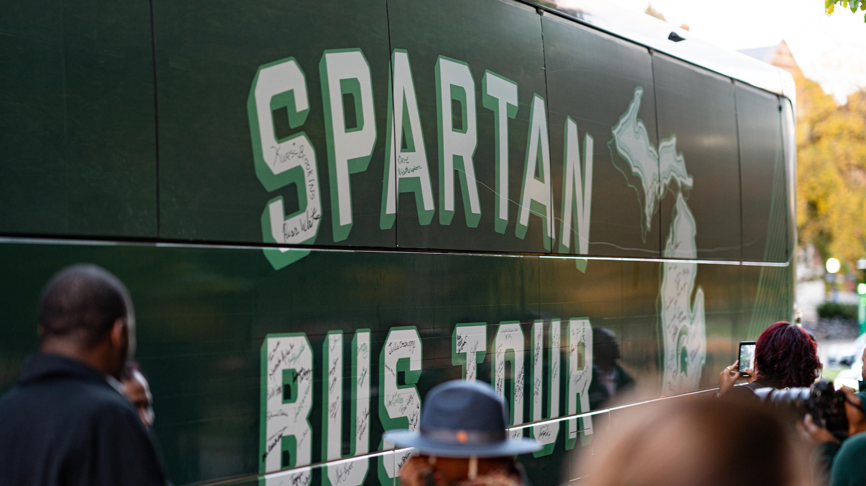 A green bus that says ?Spartan bus tour? in white with a white shape of the state of Michigan next to it. Signatures fill the letters and the outline of the state.