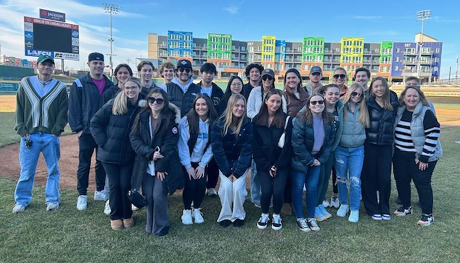 A group of people posing for a photo at a baseball field. In the background, you can see a scoreboard and apartment buildings.