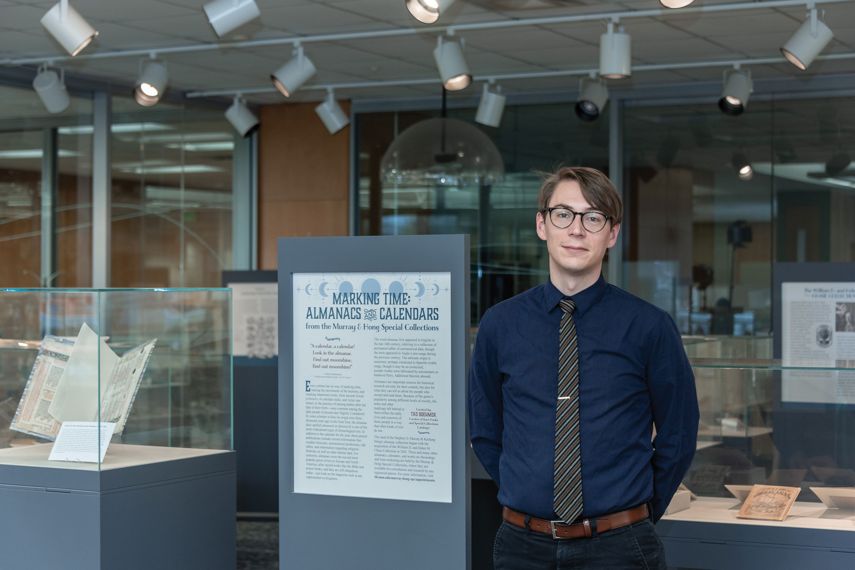 A person posing next to a sign that reads: “Marking Time: Almanacs and Calendars.” Books are displayed in glass cases behind him.