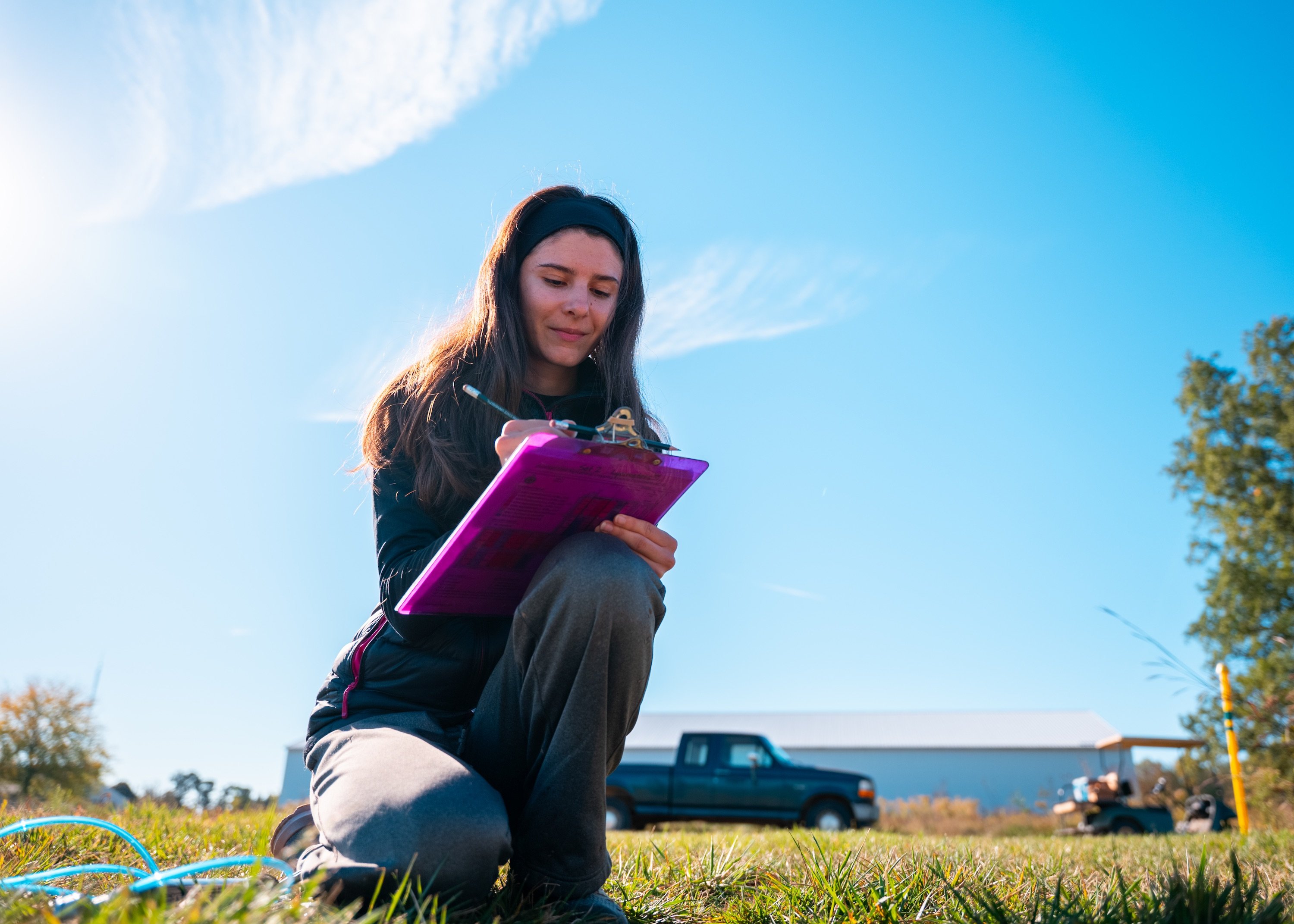 A woman writes on a purple clipboard with a truck behind her and a big expansive blue sky in the background