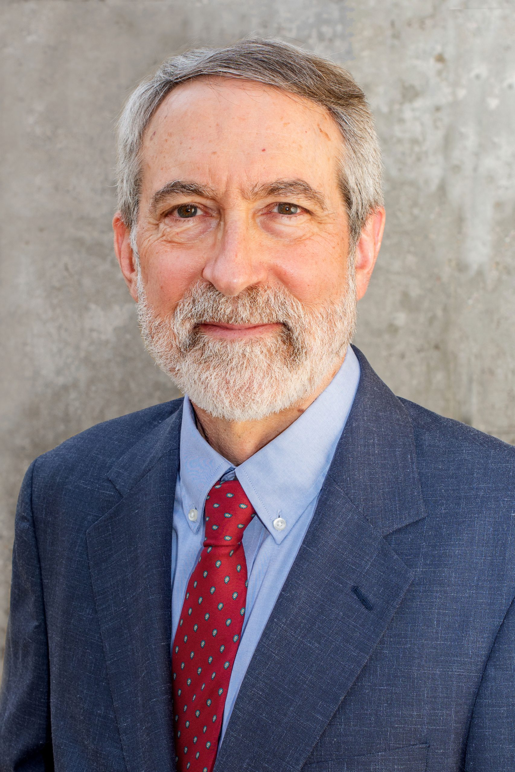 A portrait of Jeffrey Connor wearing a navy suit with a blue shirt and read tie
