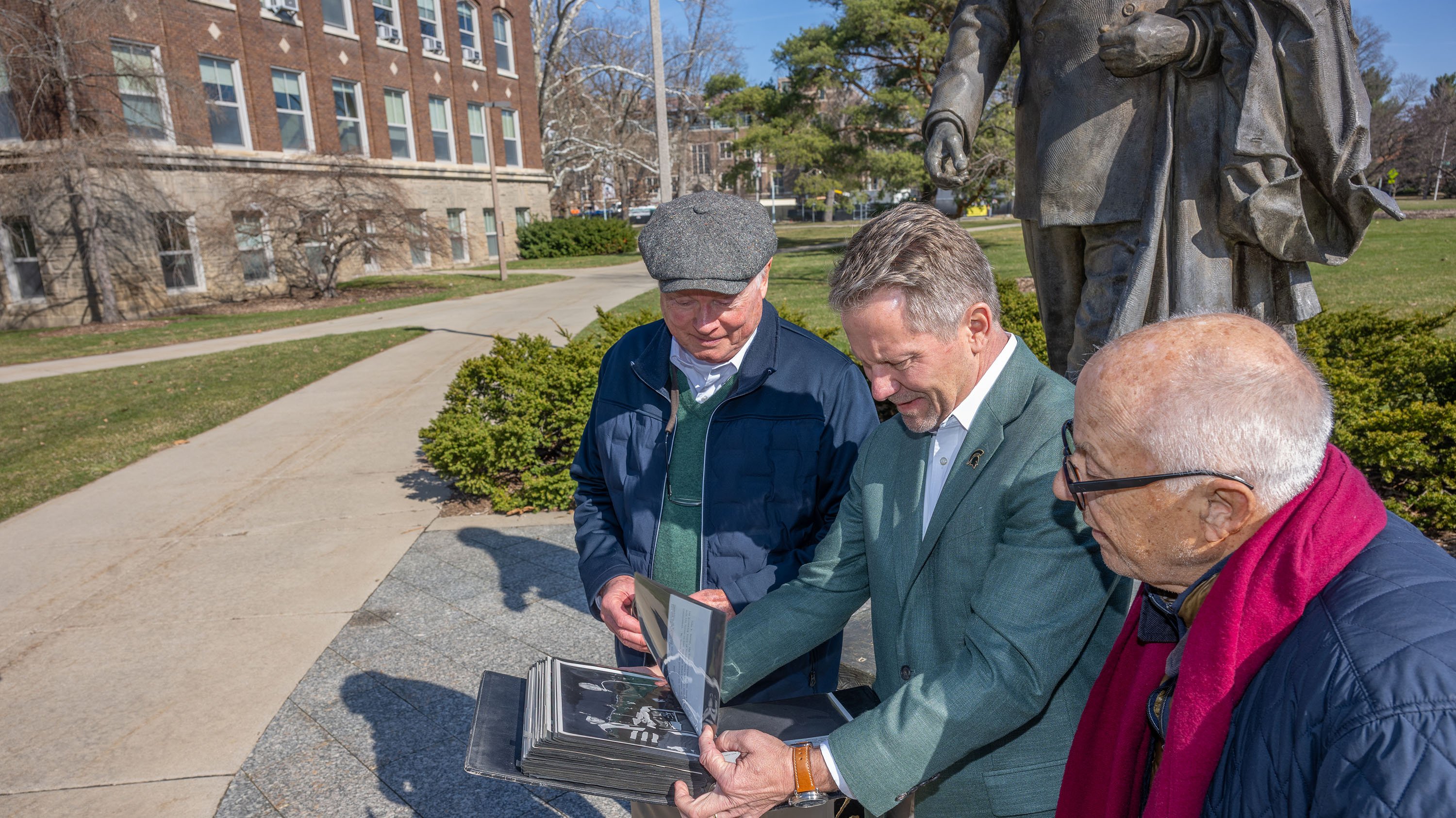 Kevin Guskiewicz, Tom Hannah--John Hannah's son, and Dirk Hornermann look at photos of former MSU President John Hannah by Hannah statue.