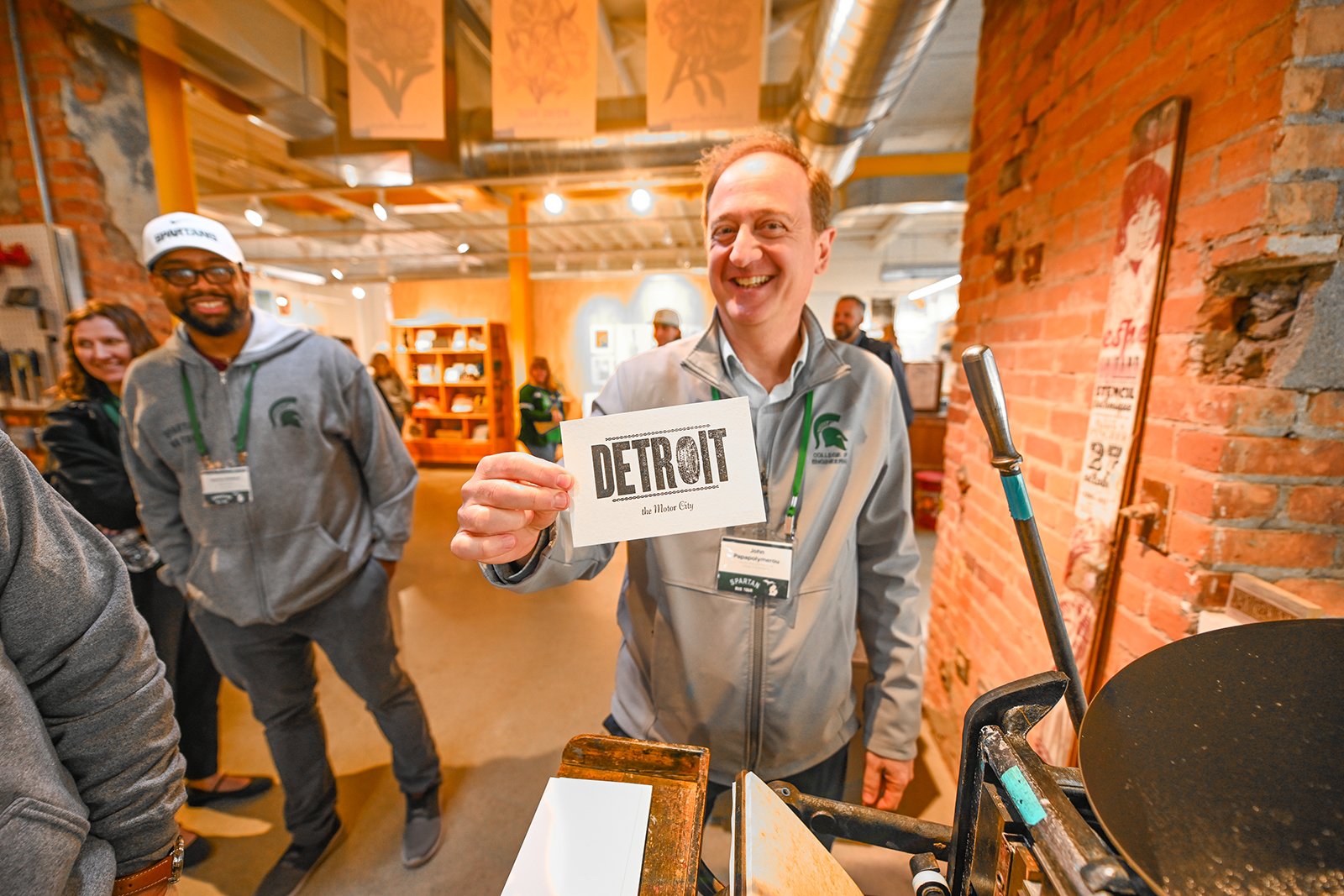 A man holds up a print that says ?Greetings from Detroit.? The ?O? in Detroit is a rubber tire.
