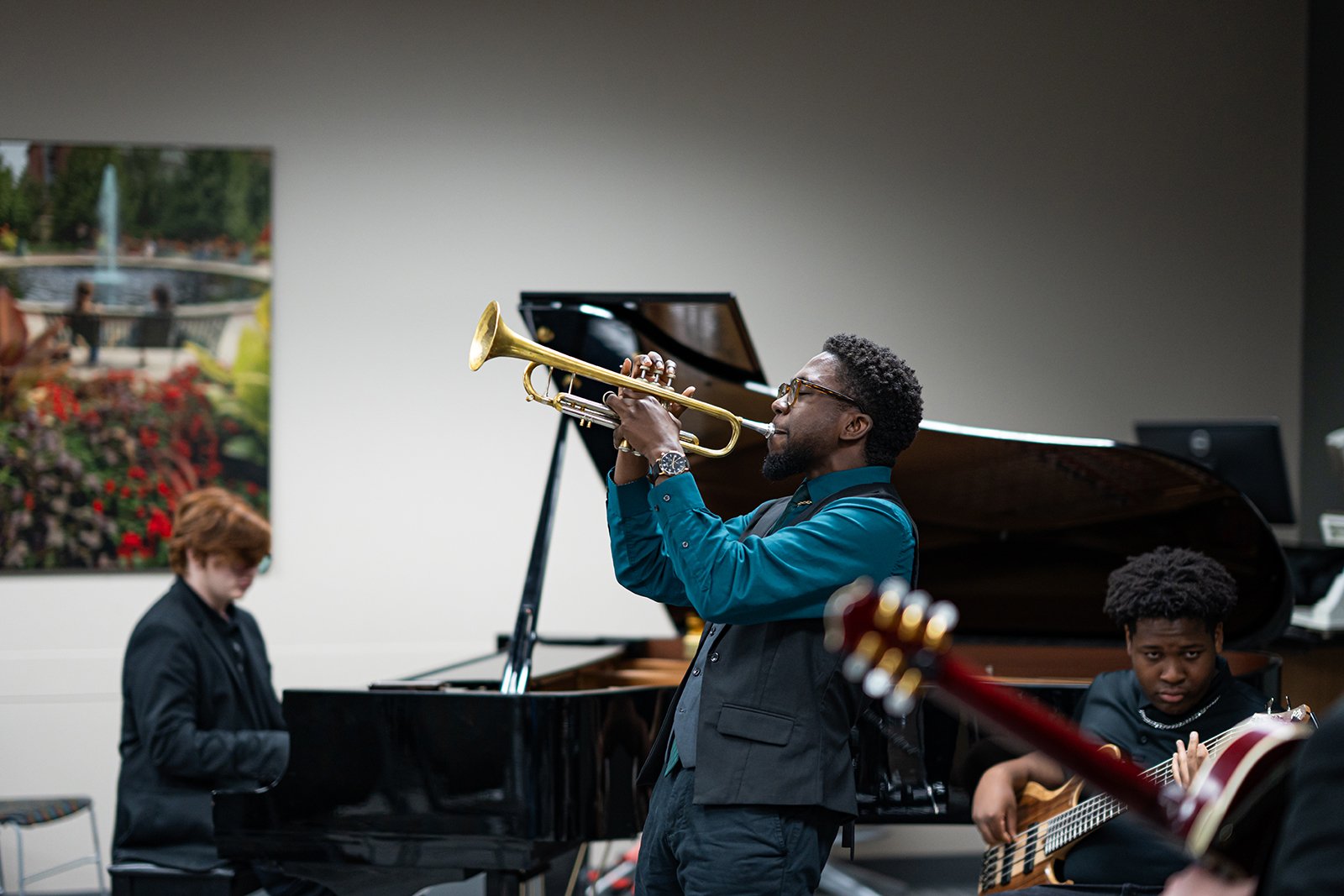 A person plays a trumpet. In the background, there is a person playing a piano.