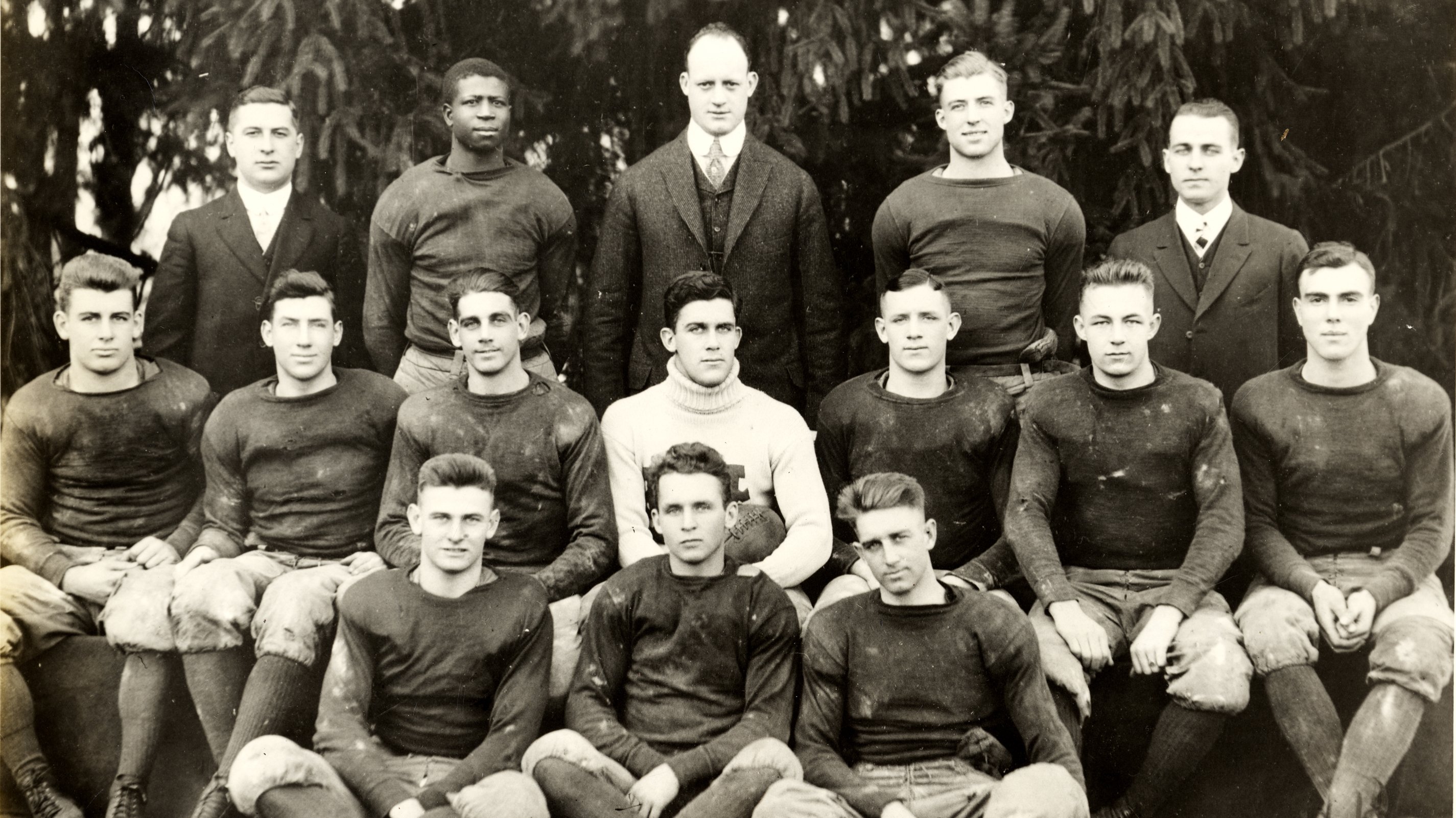A black and white photo of a football team in the early days of the sport. The players wear plain uniforms of dark long-sleeved shirts and lighter pants gathered at the knee with knee-high socks. Only one of the players is Black.