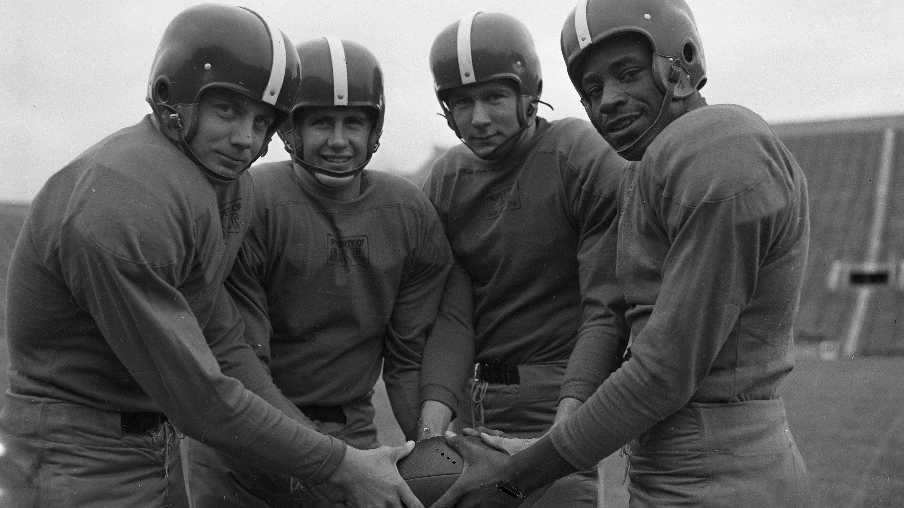 A black and white image of four football players posing for a photo. They are in a semicircle, wearing simple uniforms and helmets without facemasks, holding the same football.. One of the players is Black.