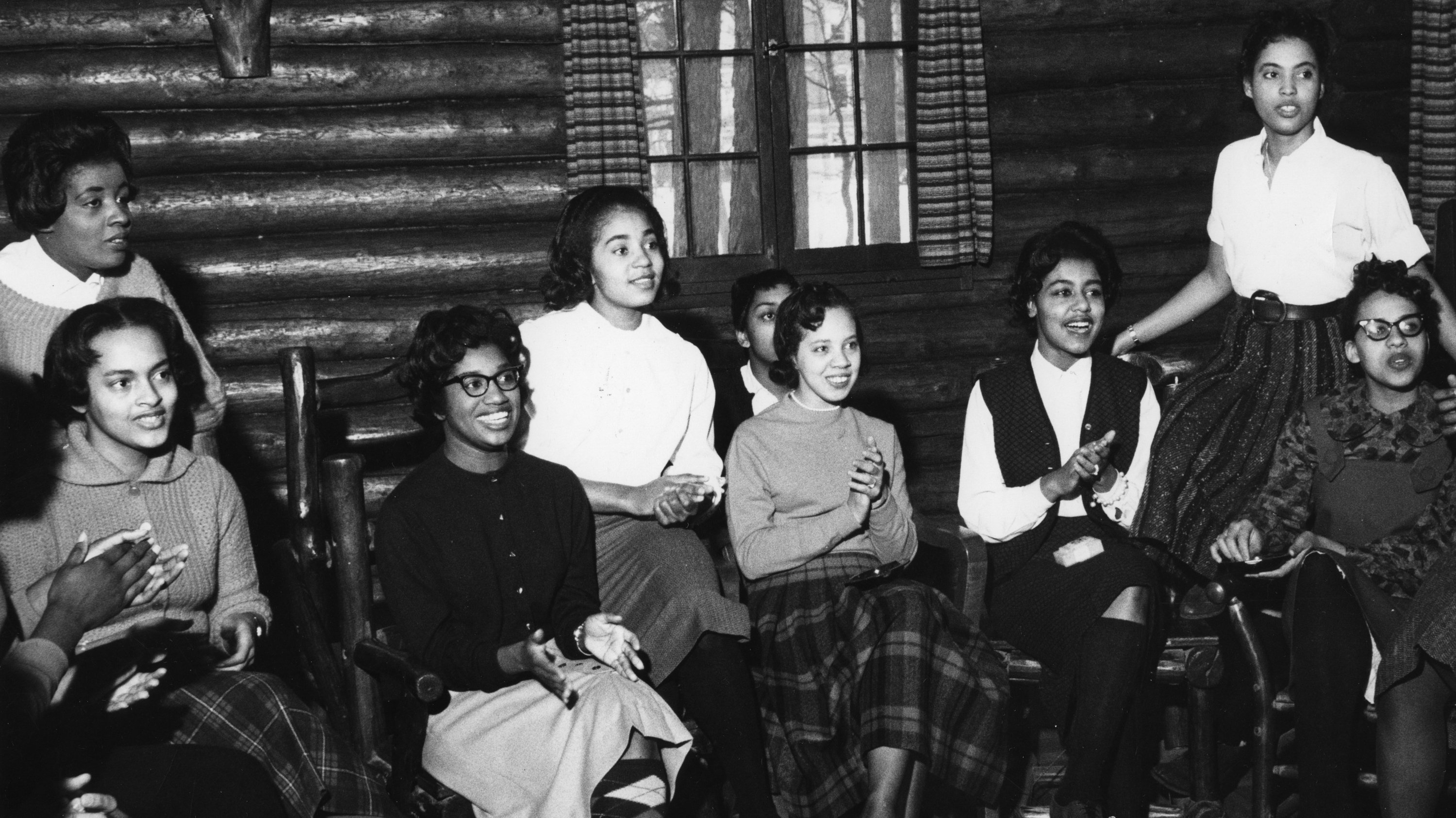 A black and white photo of nine Black women sitting on chairs in a building that resembles a log cabin. Some of the women are smiling and clapping.