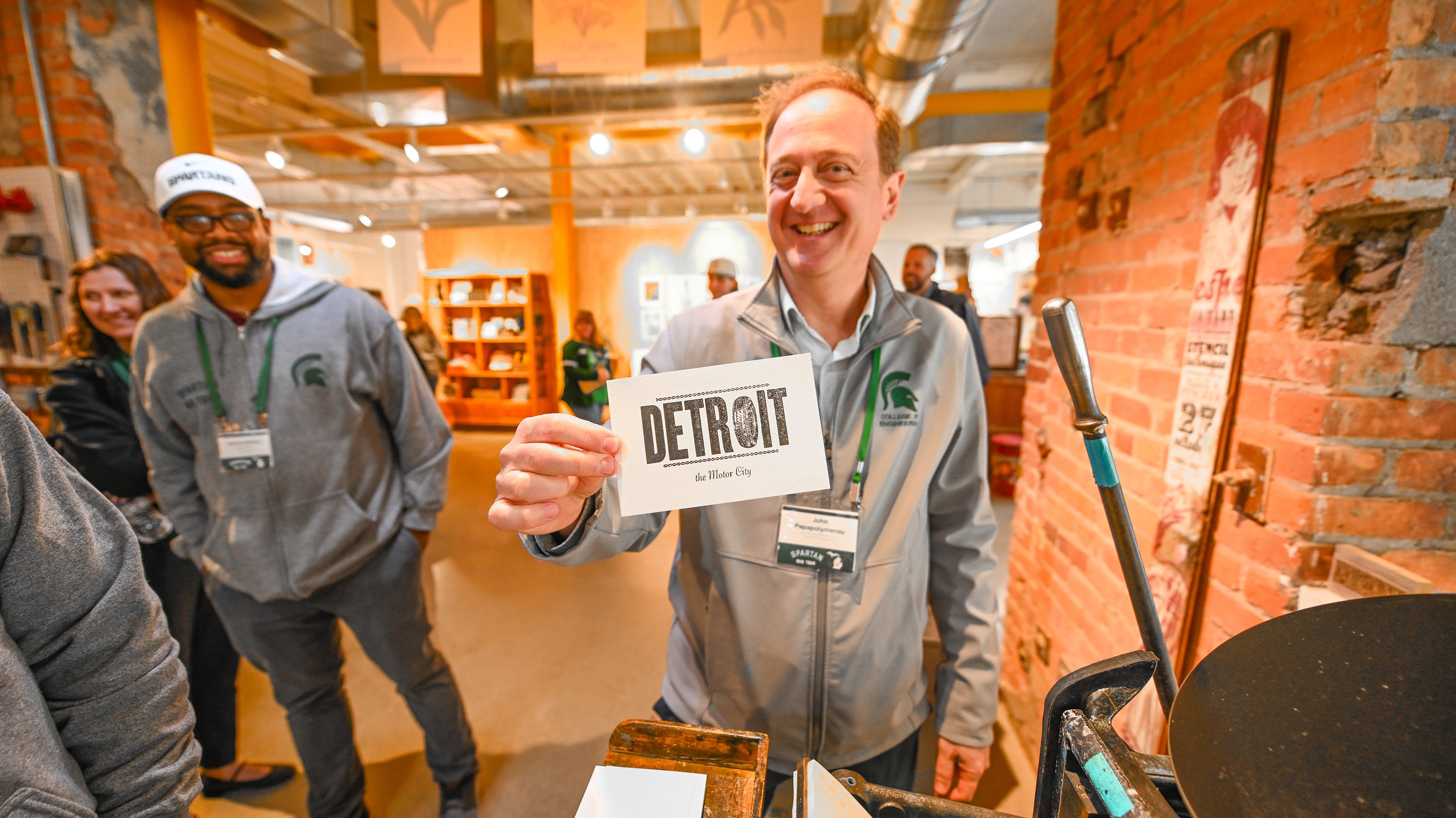 A man holds up a print that says ?Greetings from Detroit.? The ?O? in Detroit is a rubber tire.