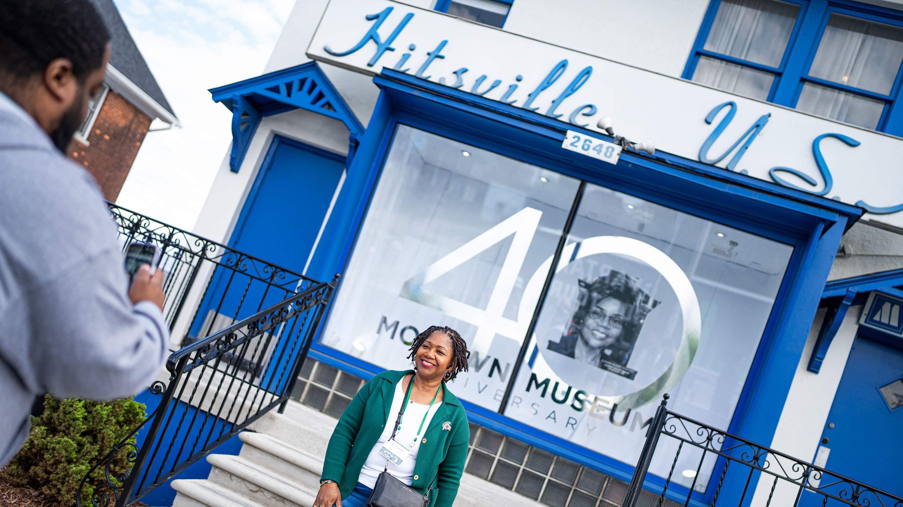 A woman poses for a photo outside of a white building with royal blue doors and trim. The words ?Hitsville USA? are on the front of the building 