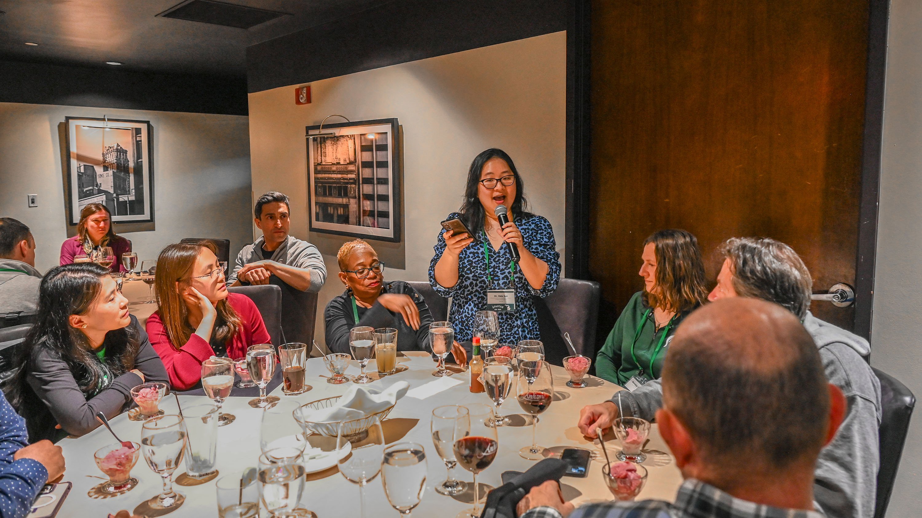 A woman holding a microphone. She is standing at a circular dinner table. Others seated at her table are looking at her.
