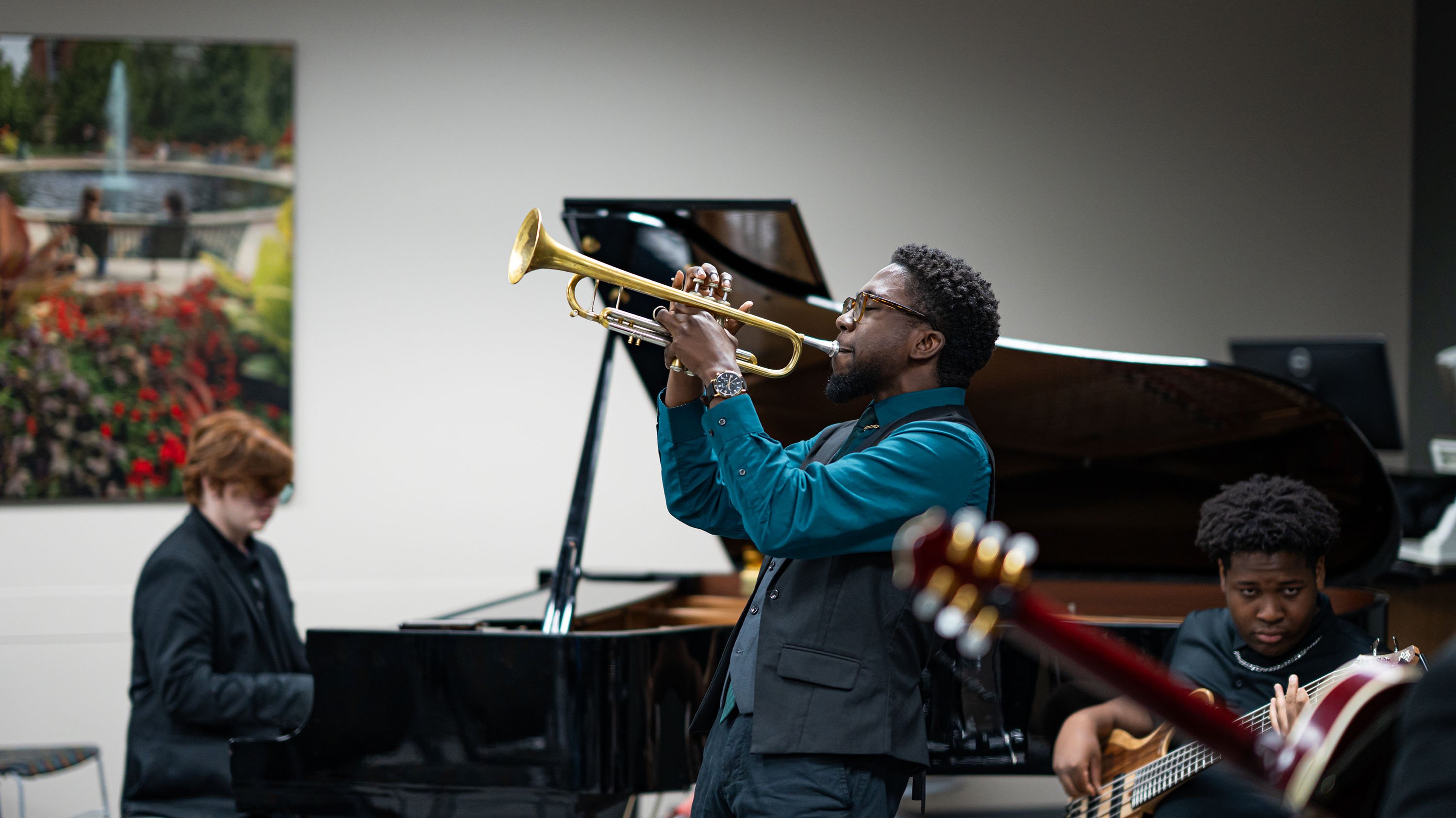 A person plays a trumpet. In the background, there is a person playing a piano.