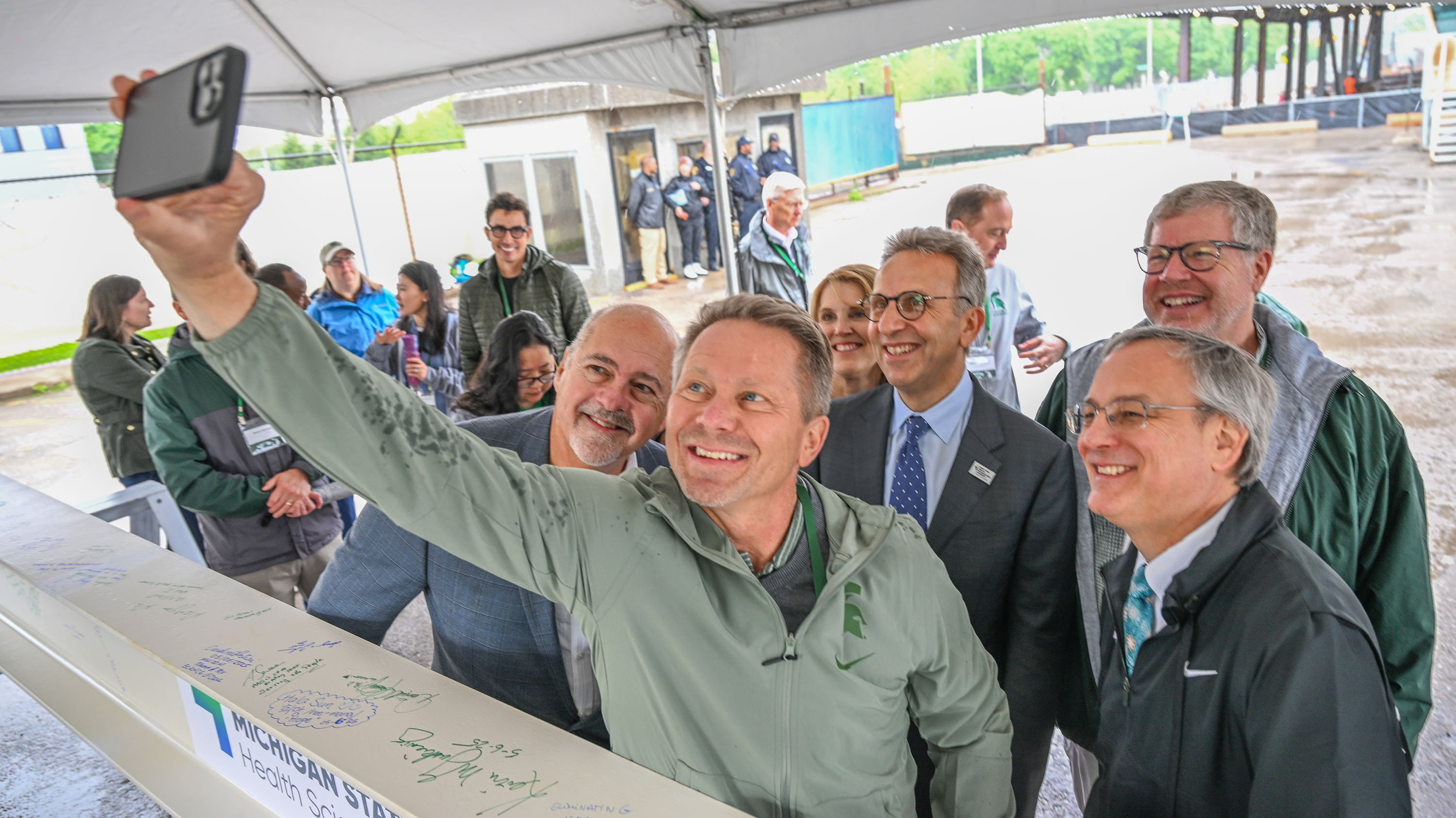 A group of people taking a selfie. They are standing near a white construction beam with signatures on it.