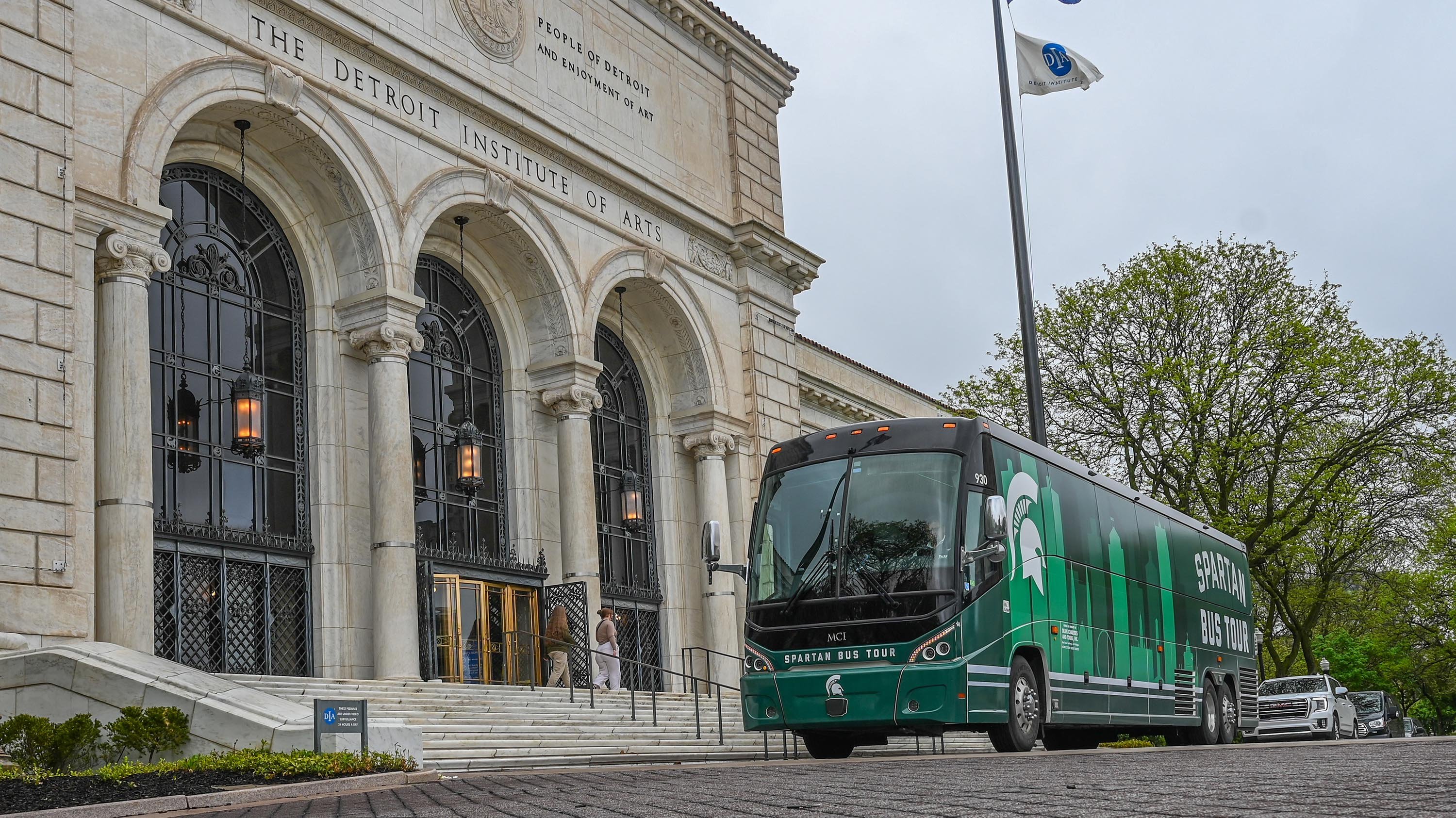 A bus with a white Spartan head and green outline of the Detroit skyline outside of a white stone building. The lettering above the entrance above the building says ?Detroit Institute of Arts?