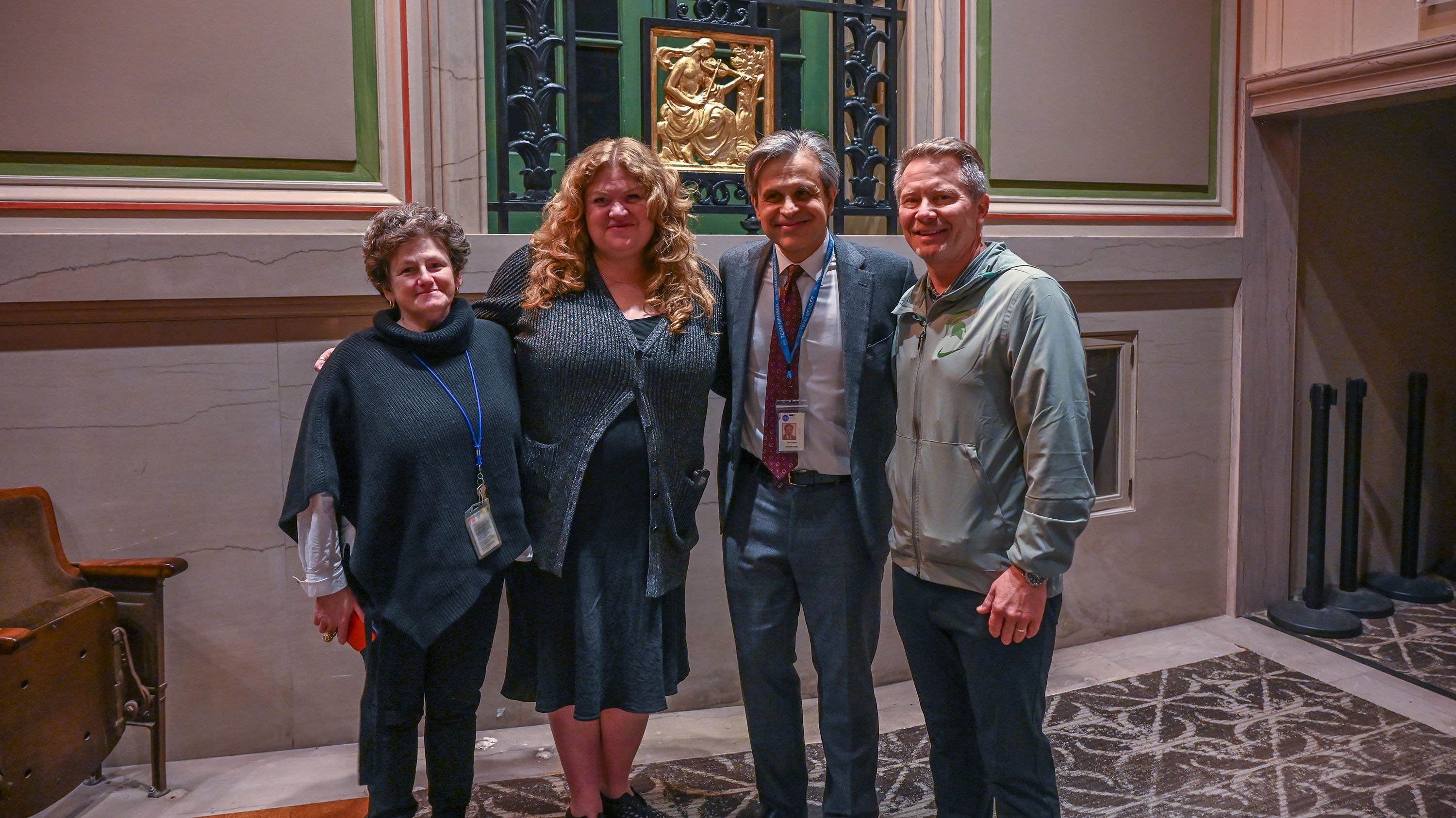 Four people pose for a photo. Behind them, there is gold square with a woman playing a string instrument with a bow carved into it.