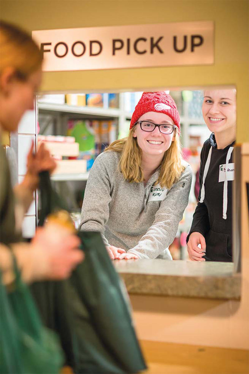 Two smiling students appear under a window that has a sign over it that reads FOOD PICK UP as another student approaches the window with a green tote bag