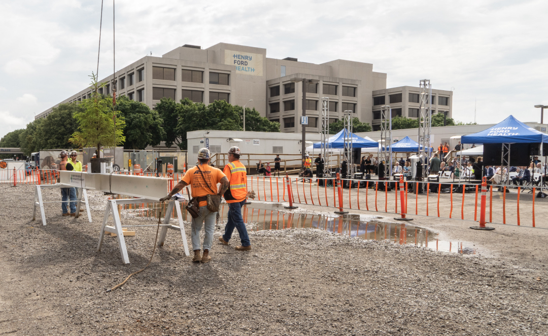 Construction workers in high-visibility vests gather around a steel beam with a small tree attached, prepared for lifting at the Henry Ford Health + MSU Research Center construction site. In the background, the Henry Ford Health building and blue tents sheltering guests at a milestone celebration event are visible.