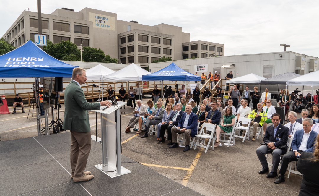 President Guskiewicz addresses a seated crowd under white and blue tents marked ?Henry Ford Health.? The audience includes formally dressed guests and construction crew members, gathered outside near the ceremonial event.