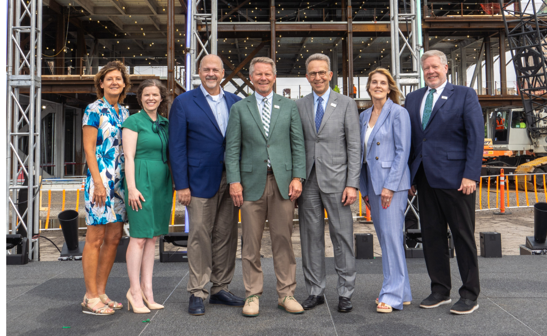 Seven leaders from Henry Ford Health and Michigan State University Health Sciences partnership pose in front of the steel framework of a new building under construction. They are standing on an outdoor stage, dressed in business and business-casual attire, smiling for a formal photo.