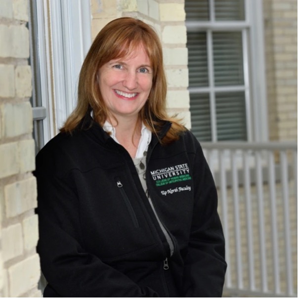 A woman with long hair smiles and leans against a brick wall wearing a black Michigan State University fleece jacket