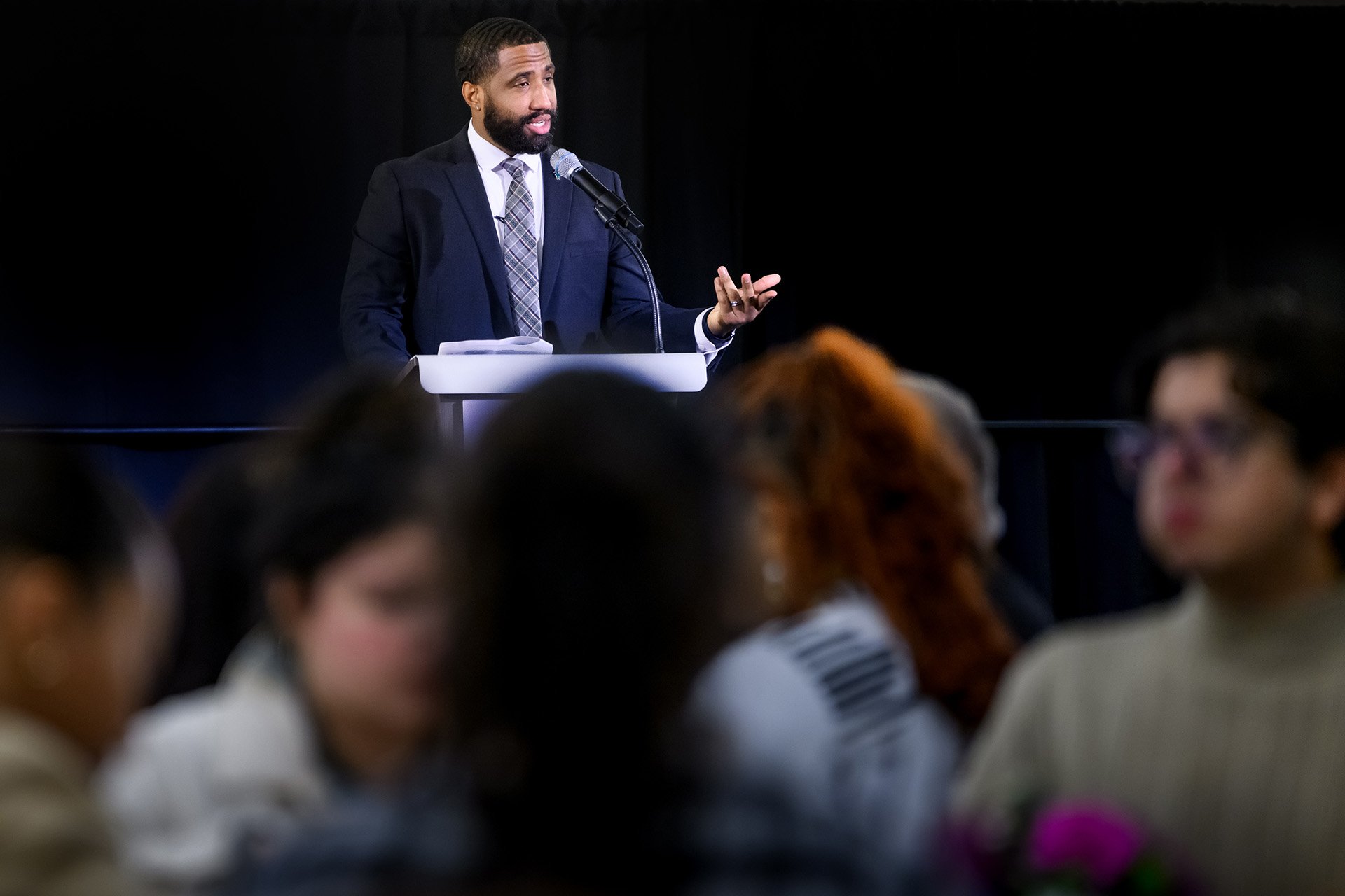 Dreux Baker welcomes the attendees standing at the lectern with audience members in the foreground
