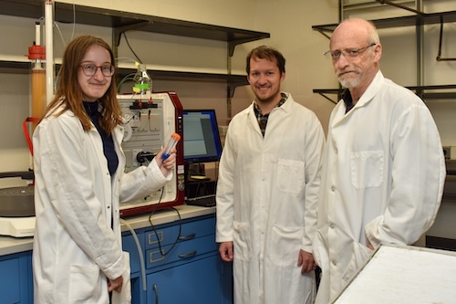 Three people in lab coats stand with a piece of scientific equipment. One of the people holds a test tube with blue liquid. Credit Kara Headley.