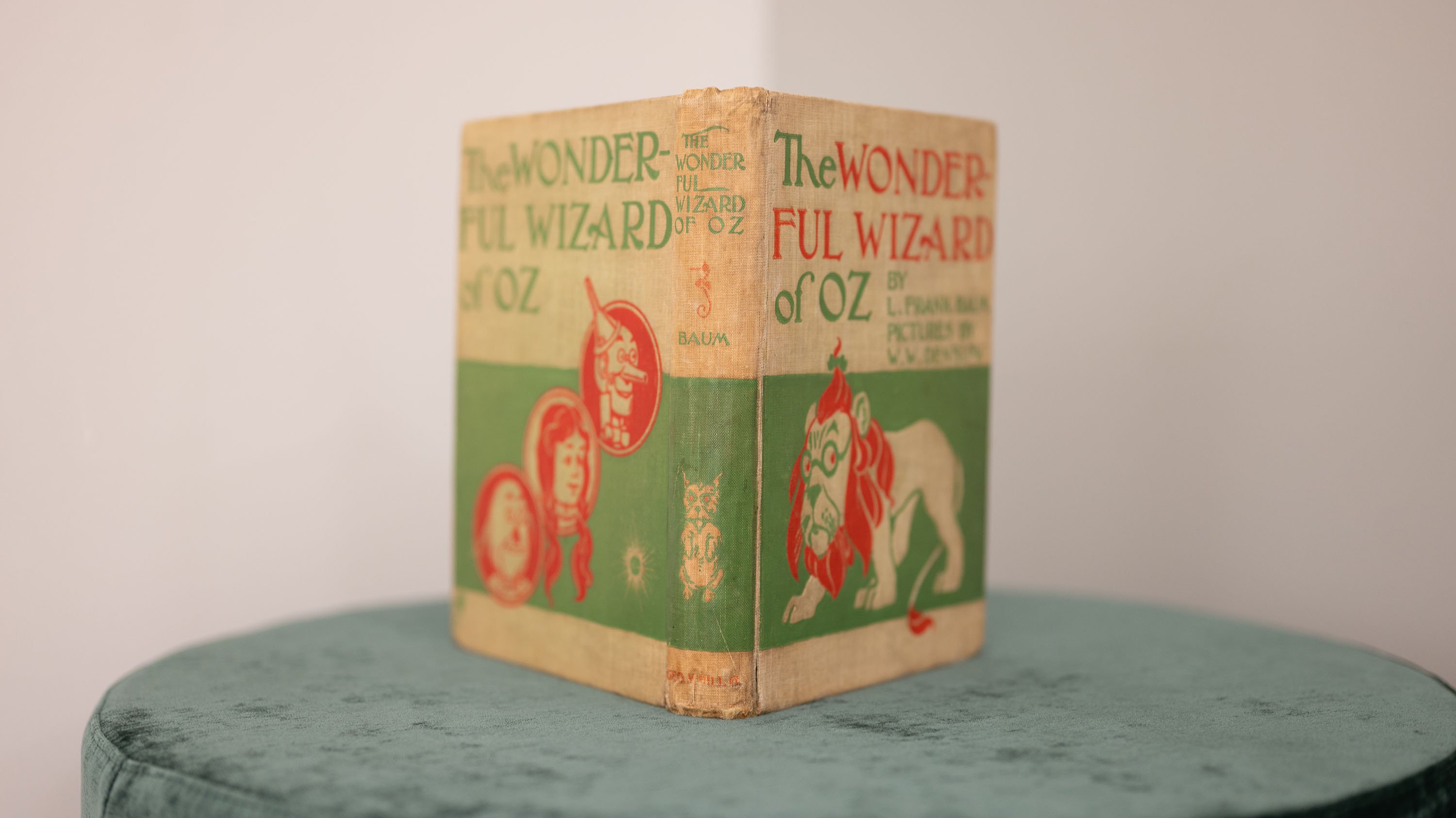 A vintage hardcover edition of The Wonderful Wizard of Oz by L. Frank Baum is displayed upright on a round, green velvet stool. The cover is beige with green and red illustrations, featuring Dorothy, the Tin Man, the Cowardly Lion, and the title in bold lettering. The book shows signs of wear along the spine and edges.