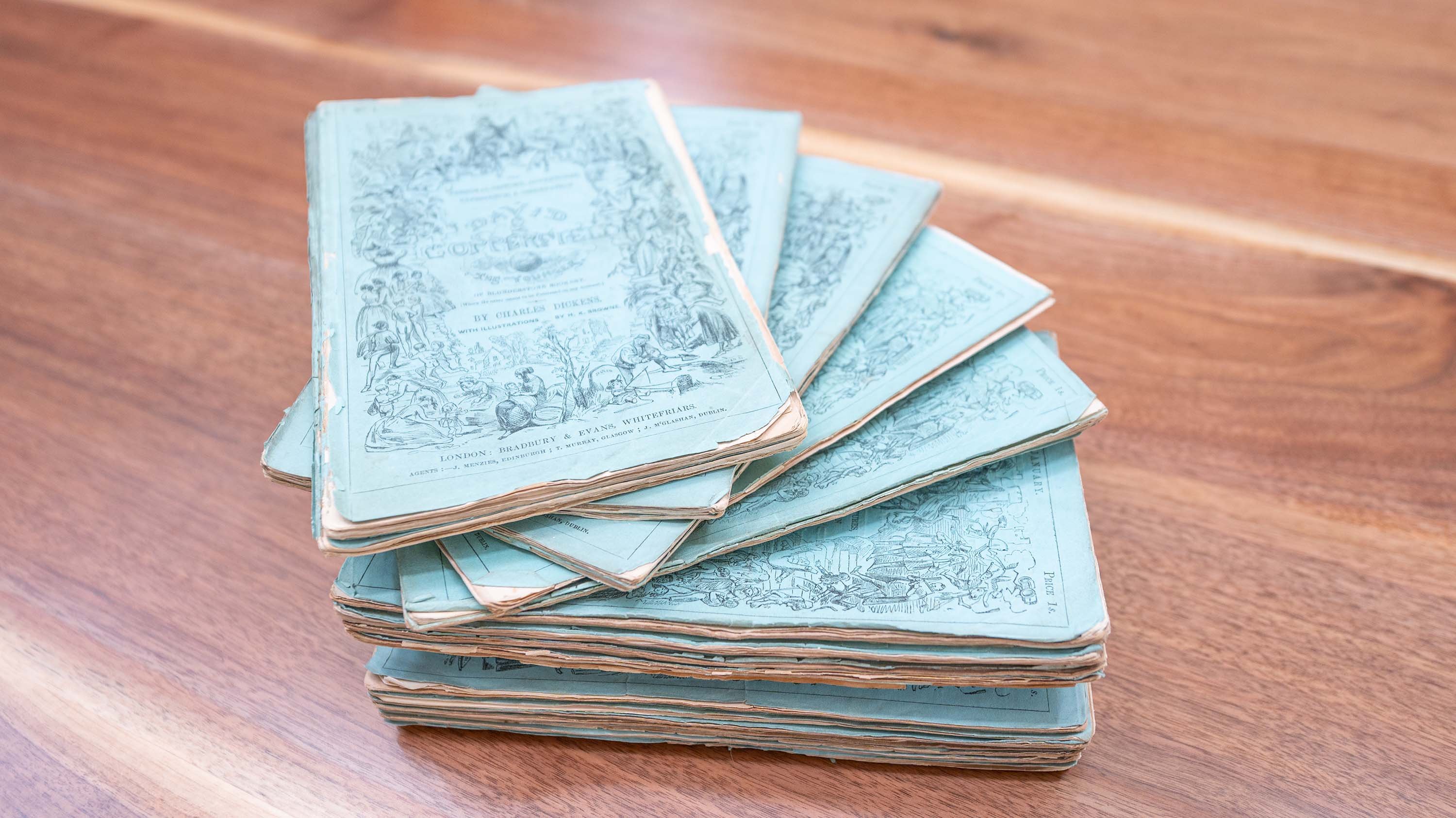 A stack of old, blue-covered periodicals of David Copperfield by Charles Dickens is fanned out on a wooden table. The covers have intricate black illustrations and text, with frayed and slightly curled edges, indicating age and frequent use.
