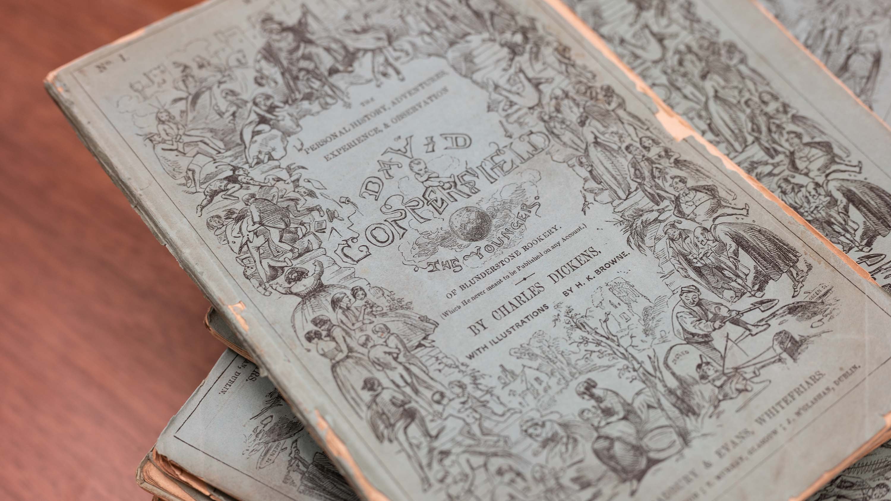 A stack of old, blue-covered periodicals of David Copperfield by Charles Dickens is fanned out on a wooden table. The covers have intricate black illustrations and text, with frayed and slightly curled edges, indicating age and frequent use.