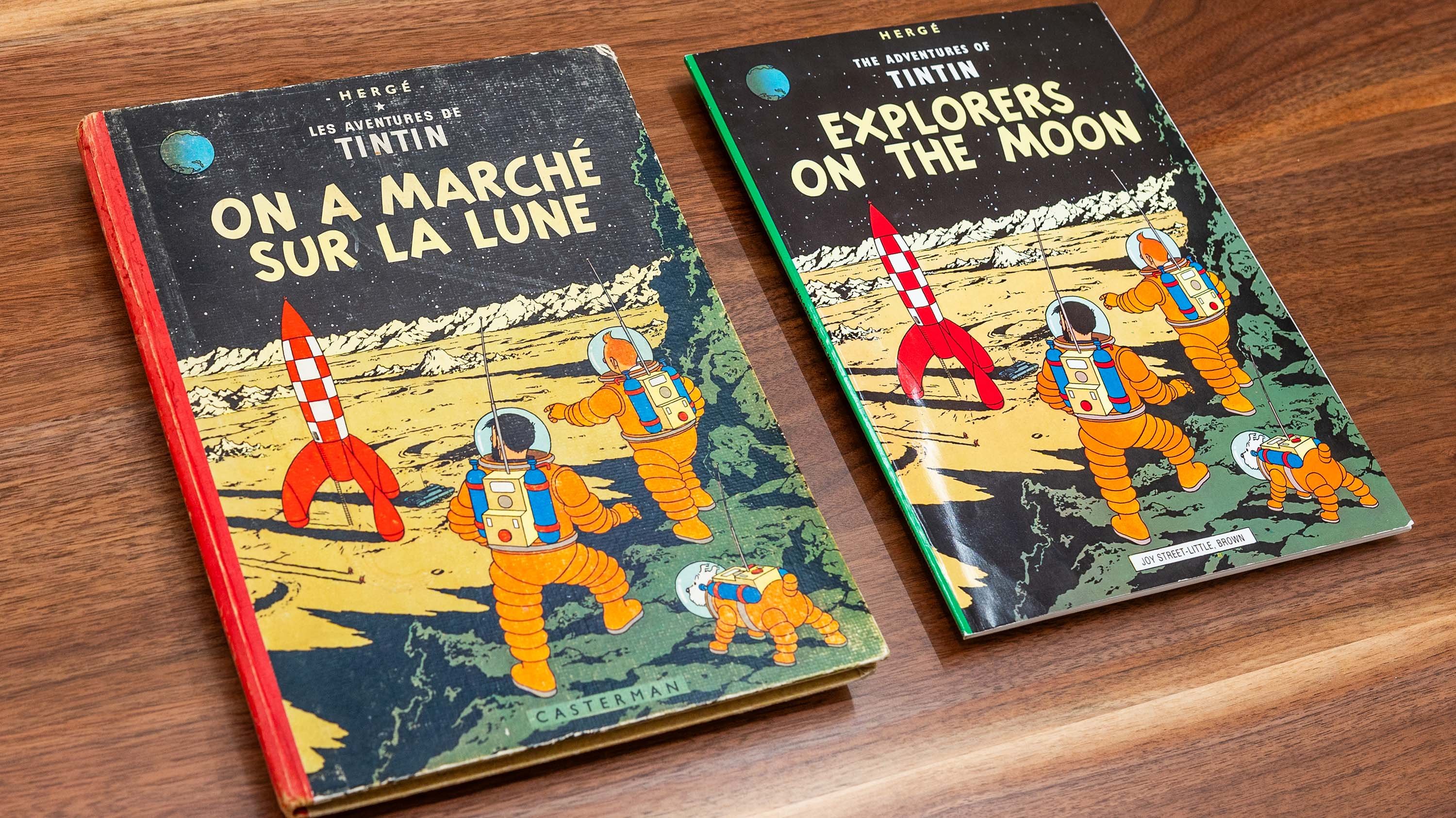 Two editions of The Adventures of Tintin: Explorers on the Moon are laid side by side on a wooden surface. One has a worn, vintage hardcover with a red spine, while the other is a newer paperback edition. Both covers feature an illustration of astronauts in orange spacesuits walking on the Moon with a red-and-white rocket in the background.