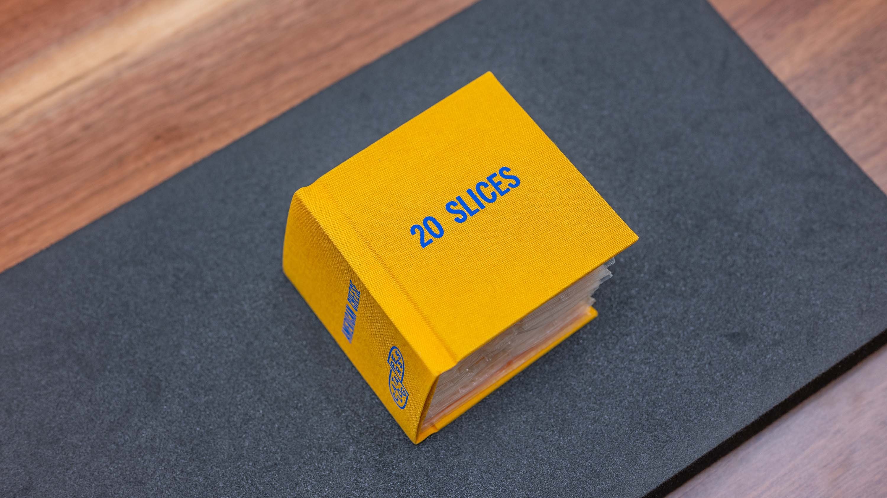 A book-shaped package made of yellow plastic, resembling a children's book. The 'cover' has the words '20 SLICES' printed in blue. Inside, you can see a stack of cheese slices wrapped in plastic.