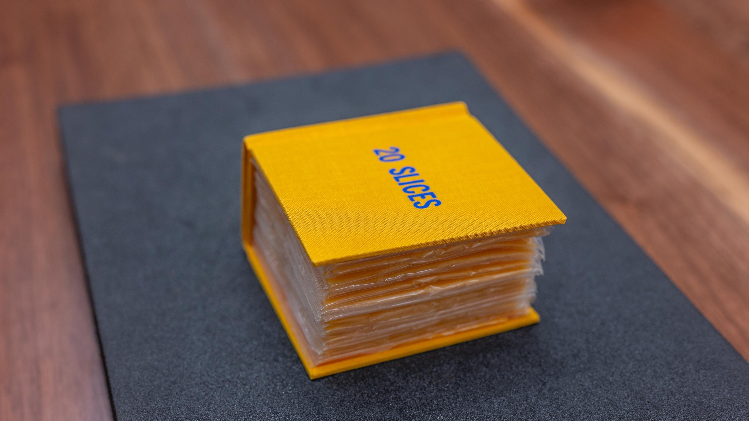 A book-shaped package made of yellow plastic, resembling a children's book. The 'cover' has the words '20 SLICES' printed in blue. Inside, you can see a stack of cheese slices wrapped in plastic.