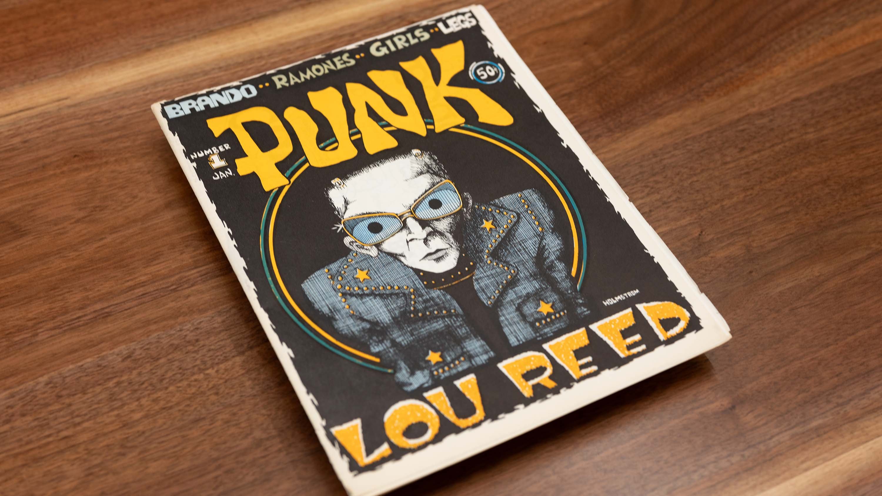A magazine titled PUNK has an illustration of a skinny man in a leather coat and glasses and in yellow font is written PUNK above the man and LOU REED beneath him