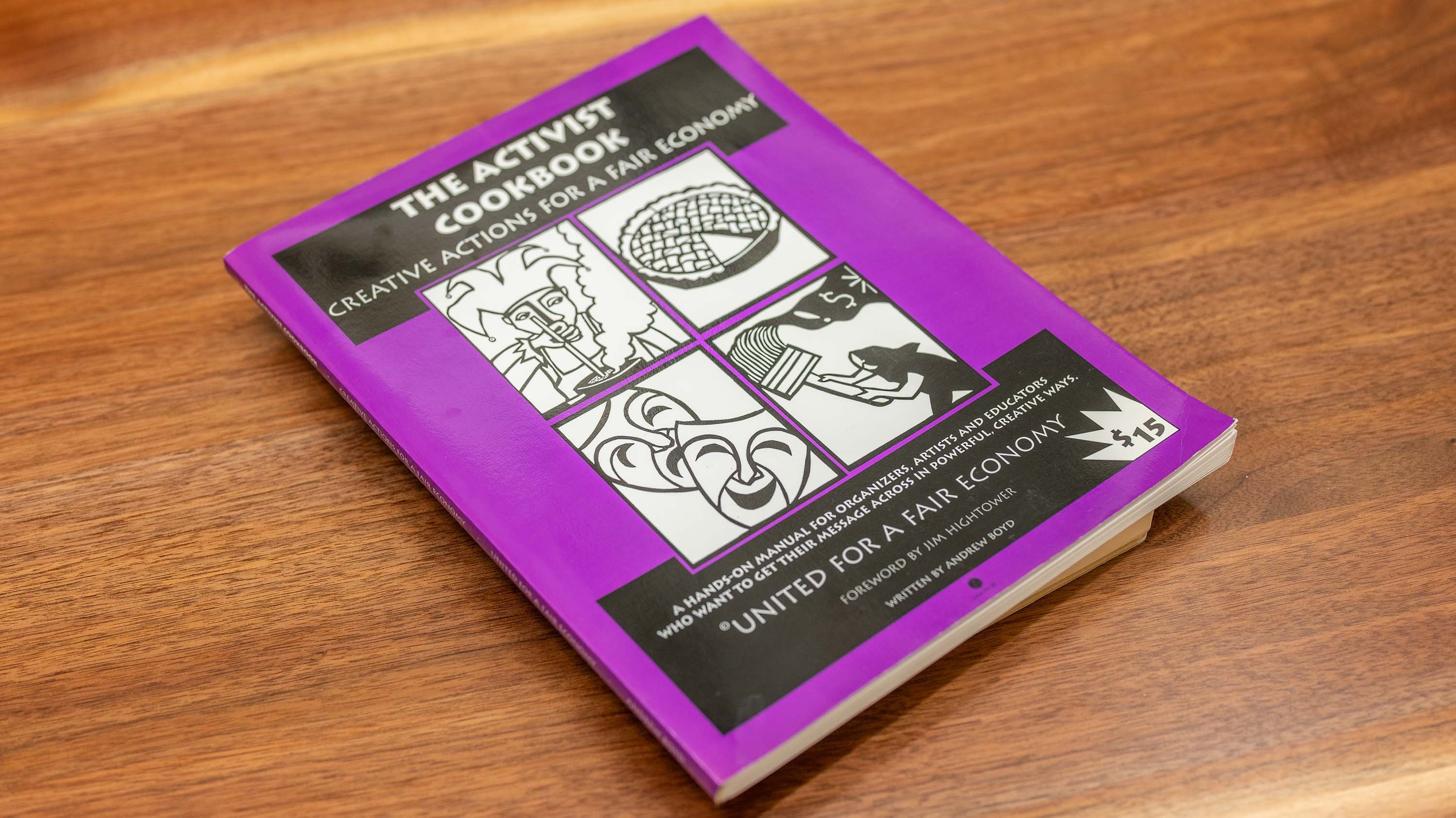 The cover of a purple book titled 'The Activist Cookbook: Creative Actions for a Fair Economy.' The cover features black and white illustrations of a pie, a paintbrush, a mask, and a megaphone.