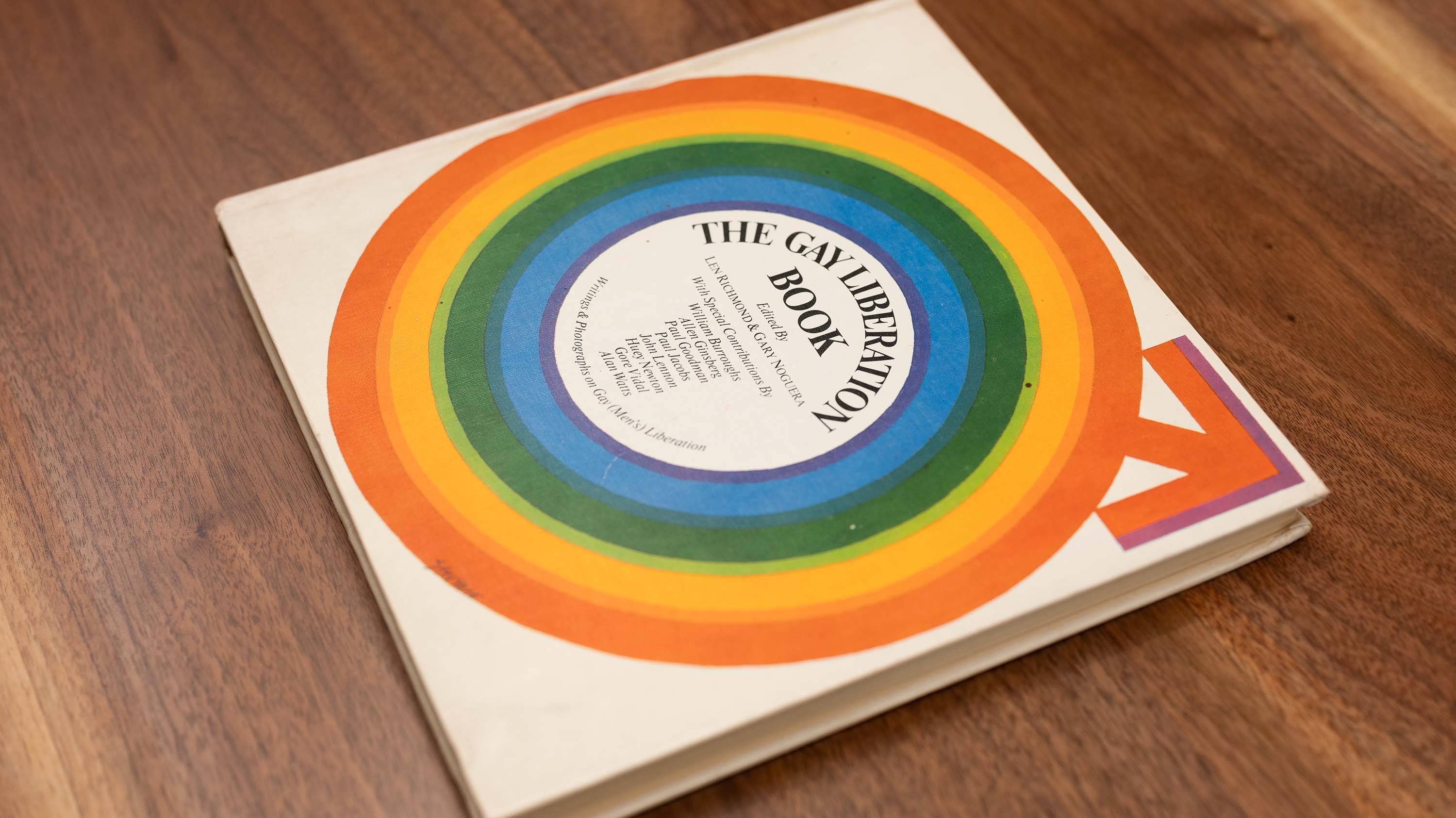 The cover of a book titled 'The Gay Liberation Book.' The cover features a rainbow circle with the title text inside. An arrow pointing to the right is below the circle.