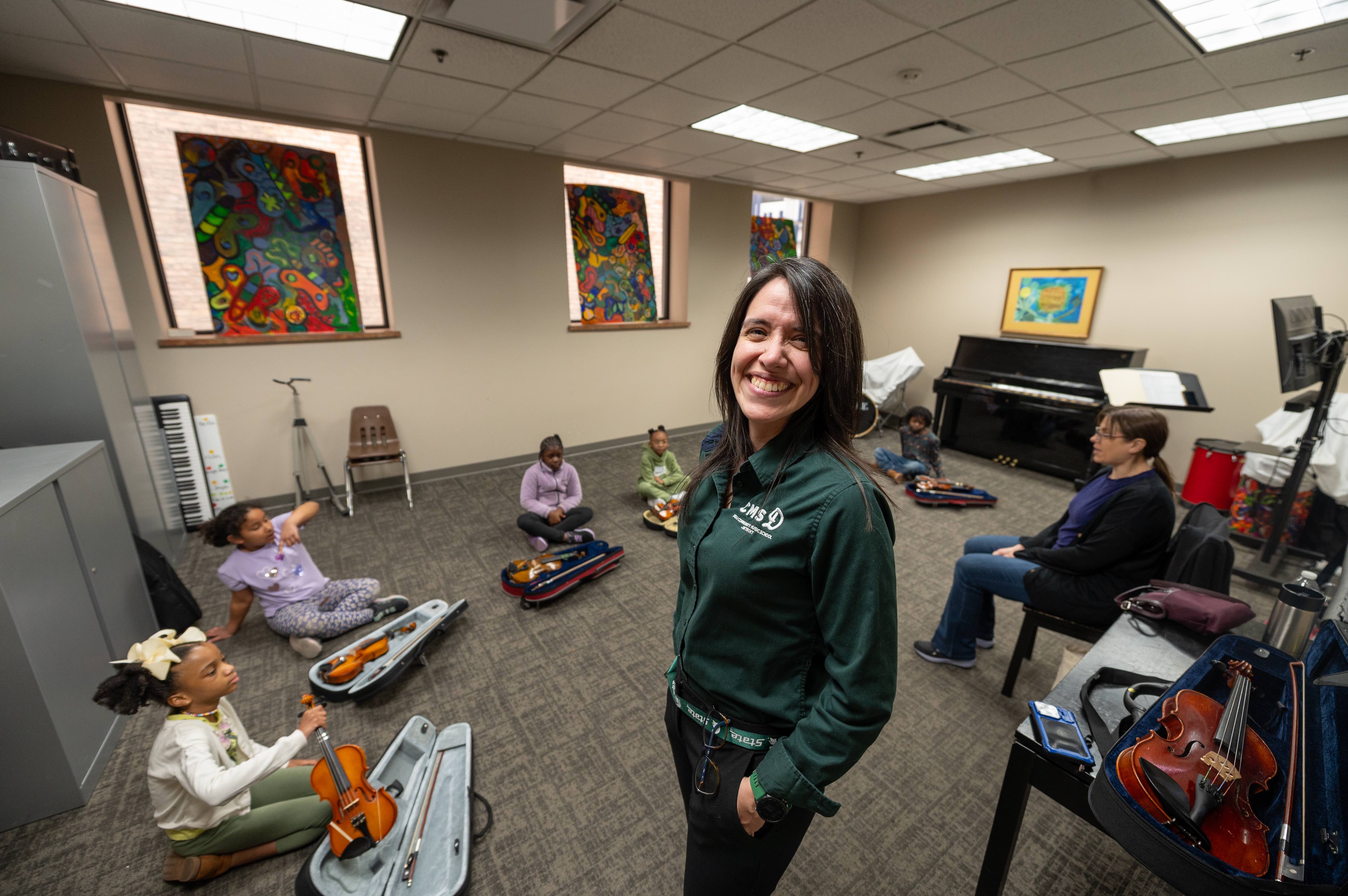 A woman in a green half-zip poses in front of a group of students sitting in a circle with instruments