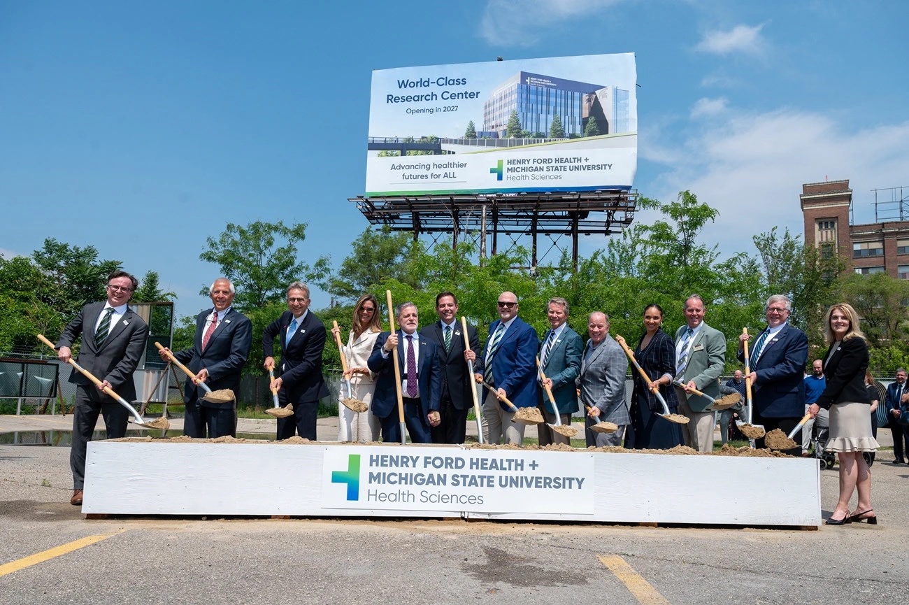 A group of people holding shovels dressed in suits poses after breaking ground in front of a sign that reads: Henry Ford Health + Michigan State University Health Sciences in front of billboard with a rendering of a new hospital building