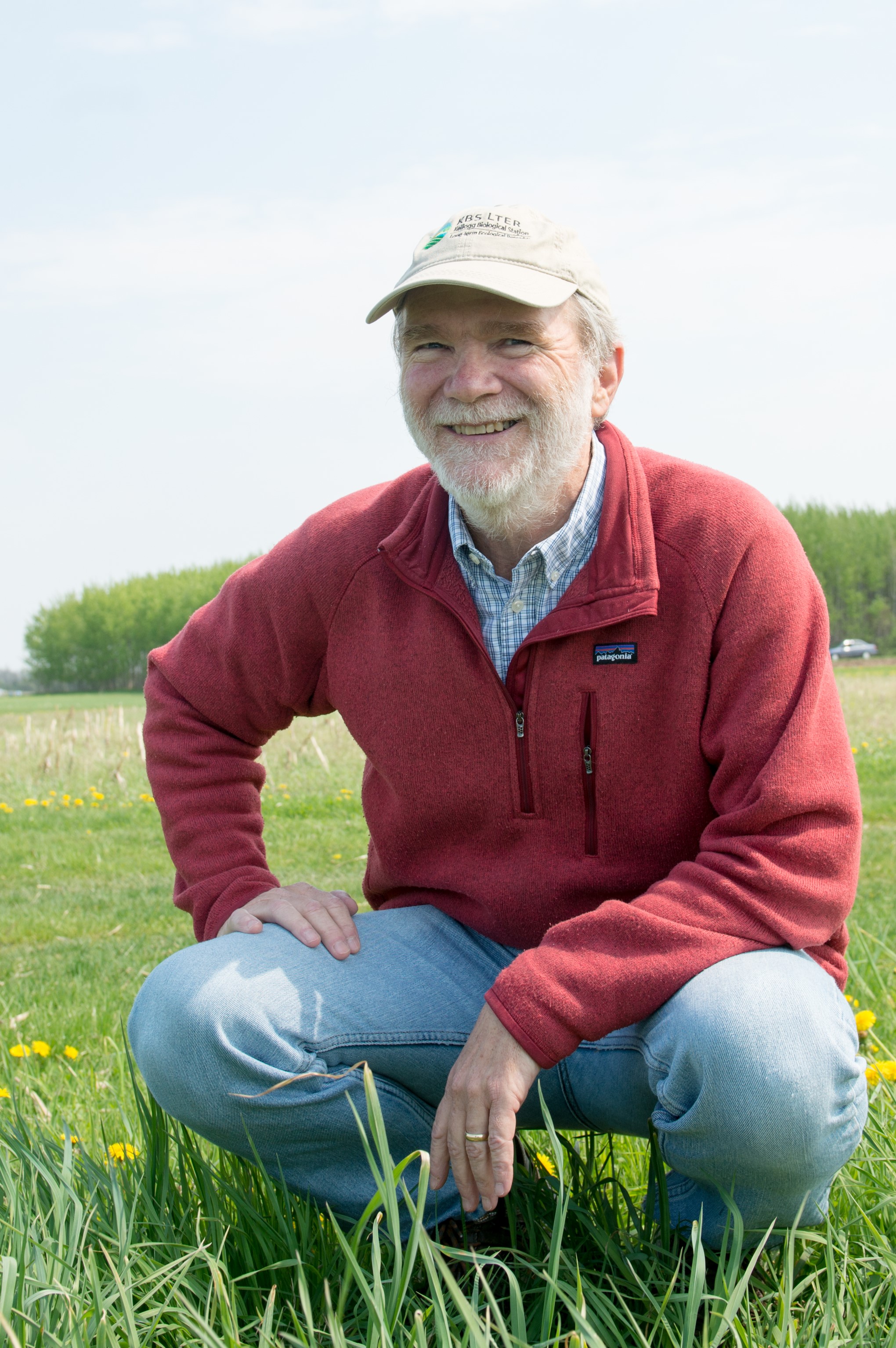 Man with white beard kneels in a field wearing jeans, a red sweater and a hat