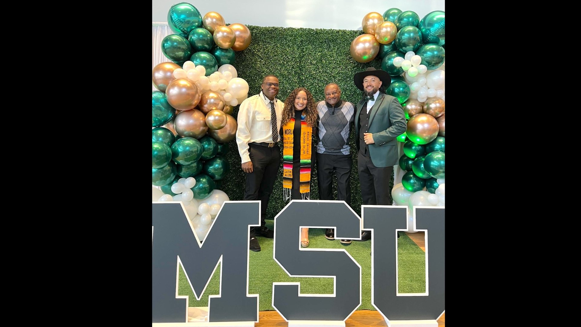 a group stands with a student wearing graduation gown in front of a backdrop