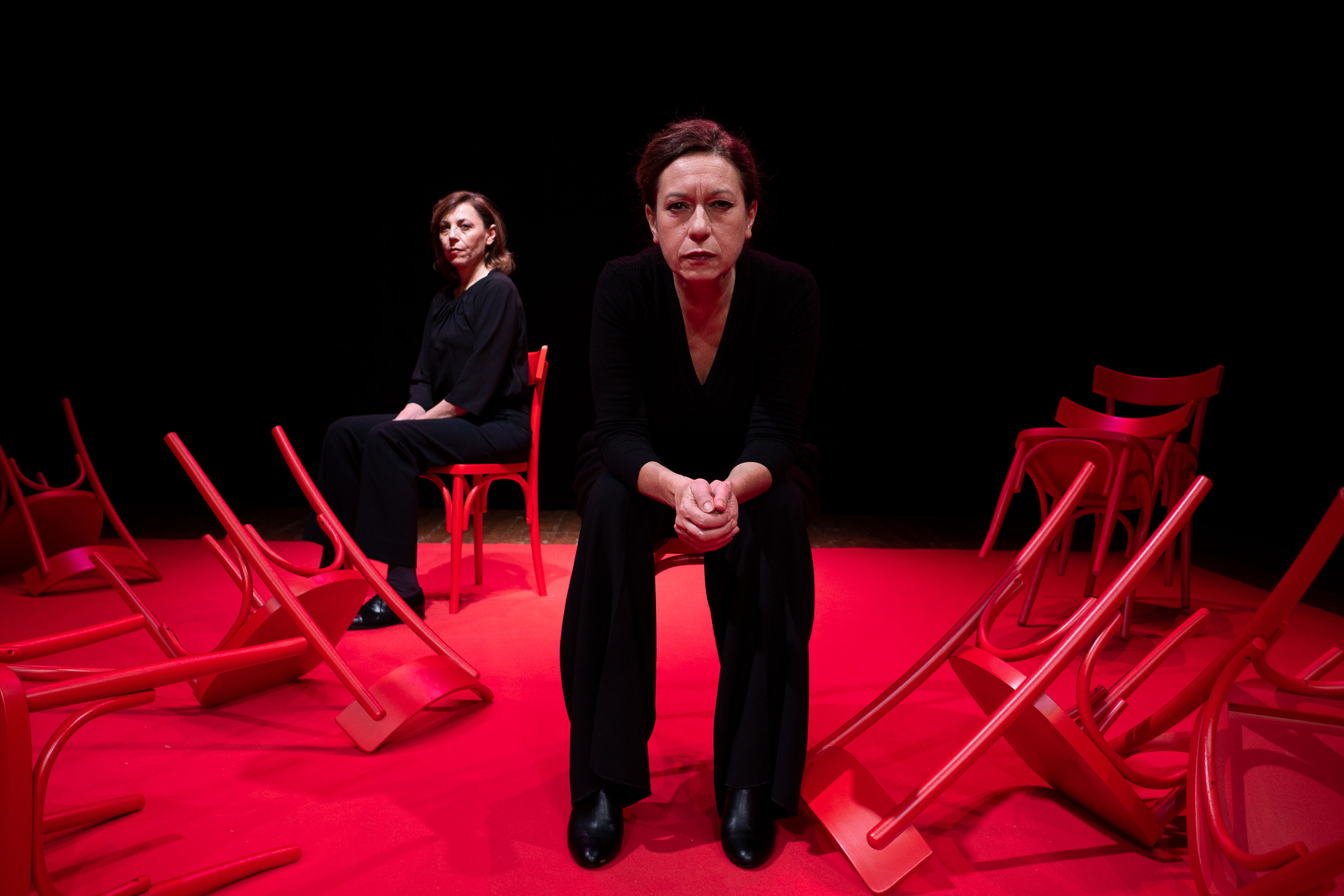 wo women wearing black sit on a theatre stage that is lit in red. One woman is sitting and looking straight ahead. The other woman is more in the background of the left and sitting at an angle. A group of discarded chairs surround both women.